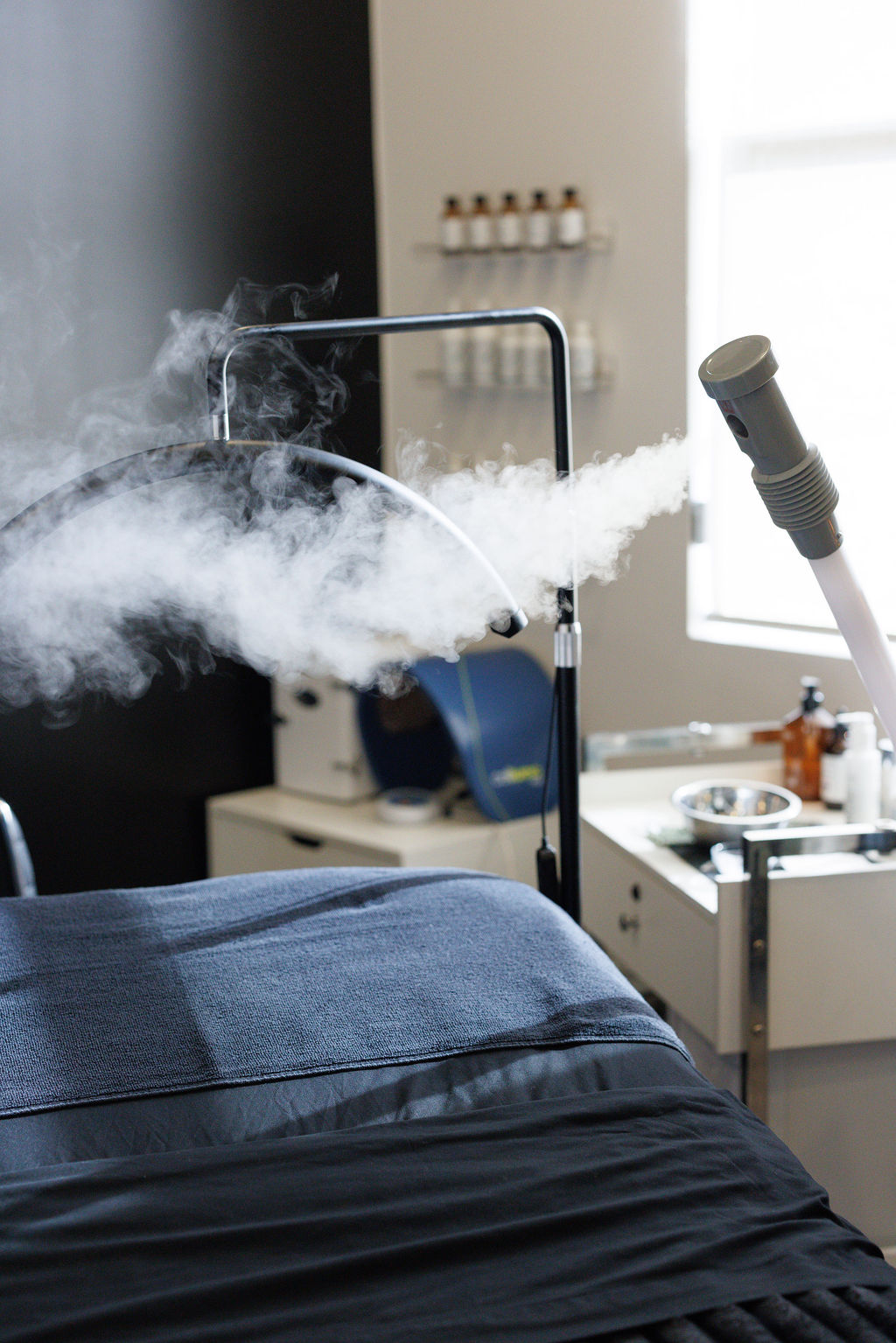 Medical equipment with vapor or mist near a bed in a clinical setting, with a white wall and shelves in the background.