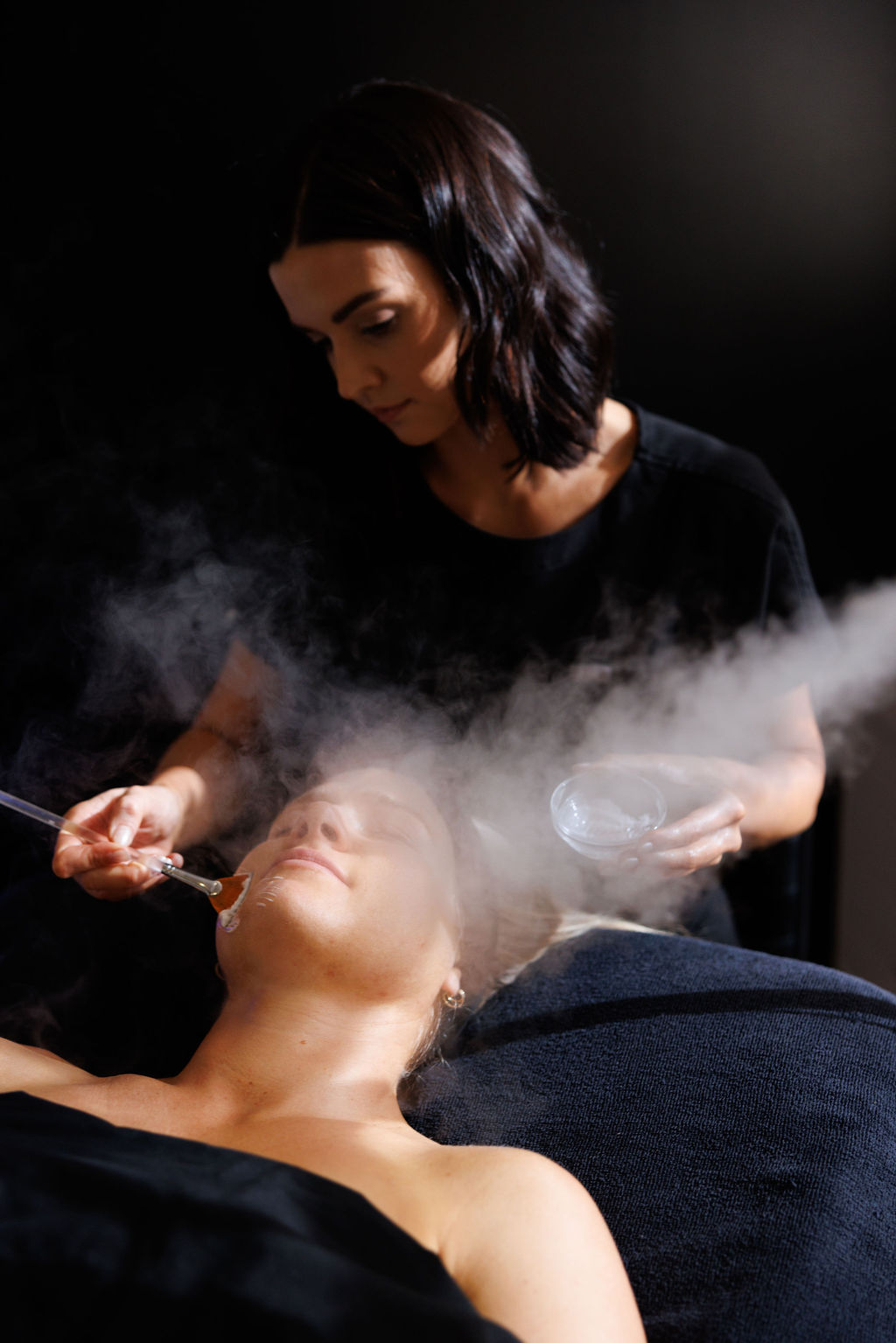 A woman performs a facial treatment on a person lying down, with steam rising from the face, in a dark room.