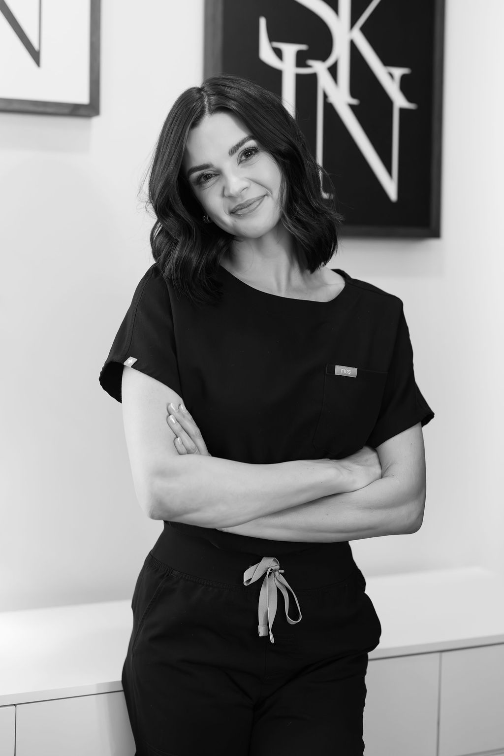 Young woman with shoulder-length hair smiling, arms crossed, wearing a black t-shirt and pants, standing indoors.
