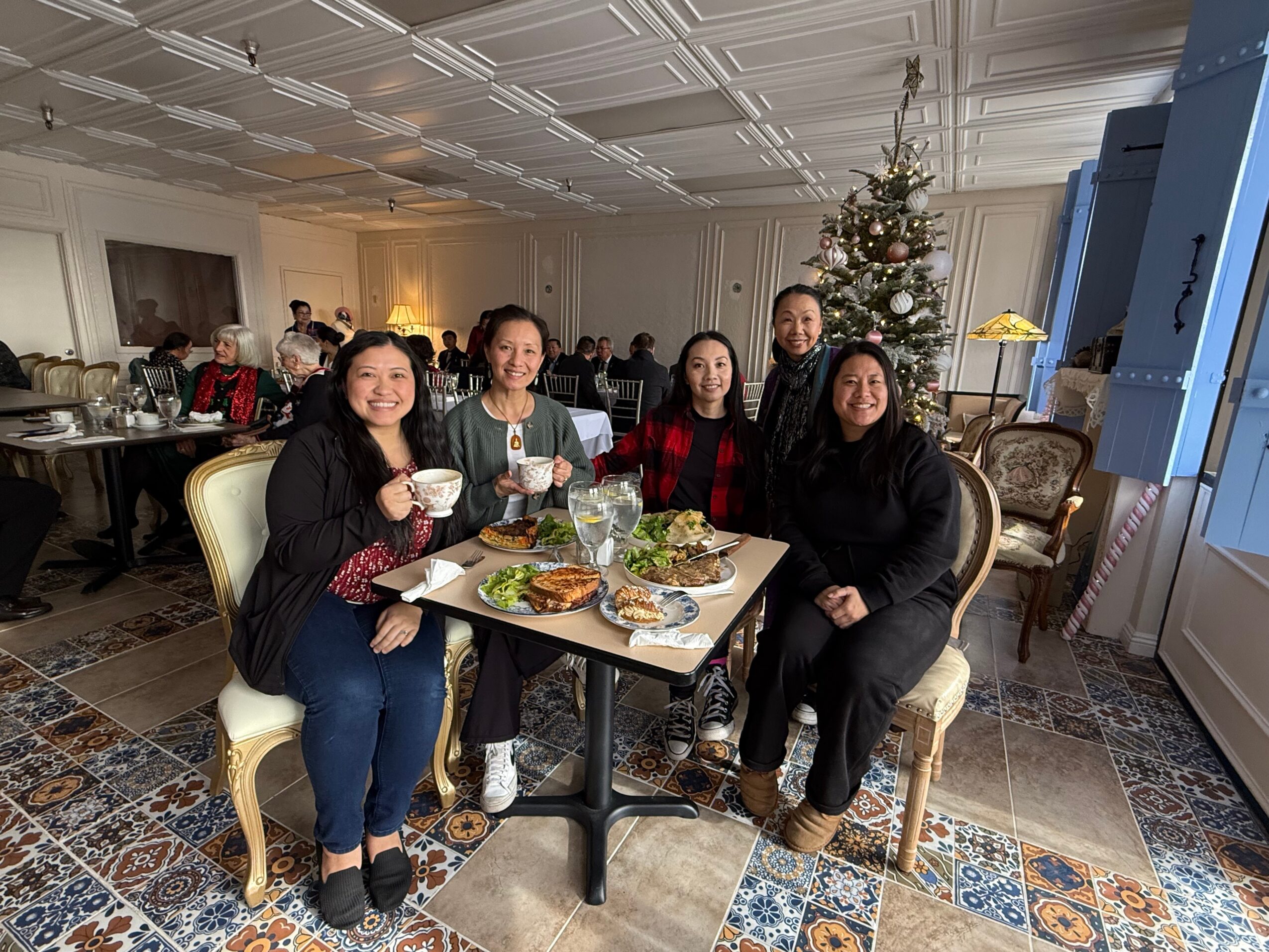 Five people sit around a table with food and drinks, smiling at a festive event with a decorated Christmas tree in the background.