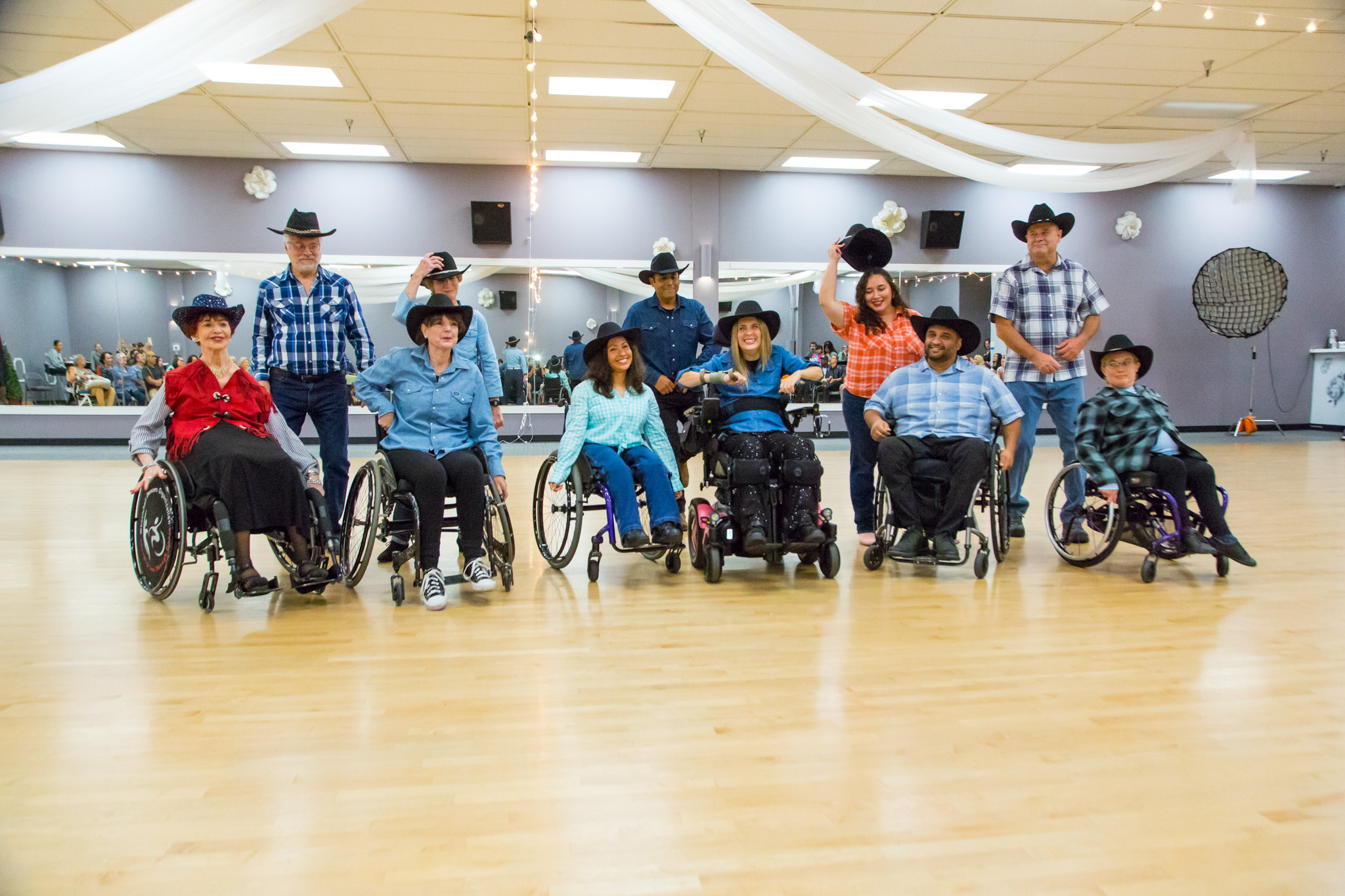 Group of people, some in wheelchairs, standing in a spacious room with wooden floor and decorative ceiling drapes.