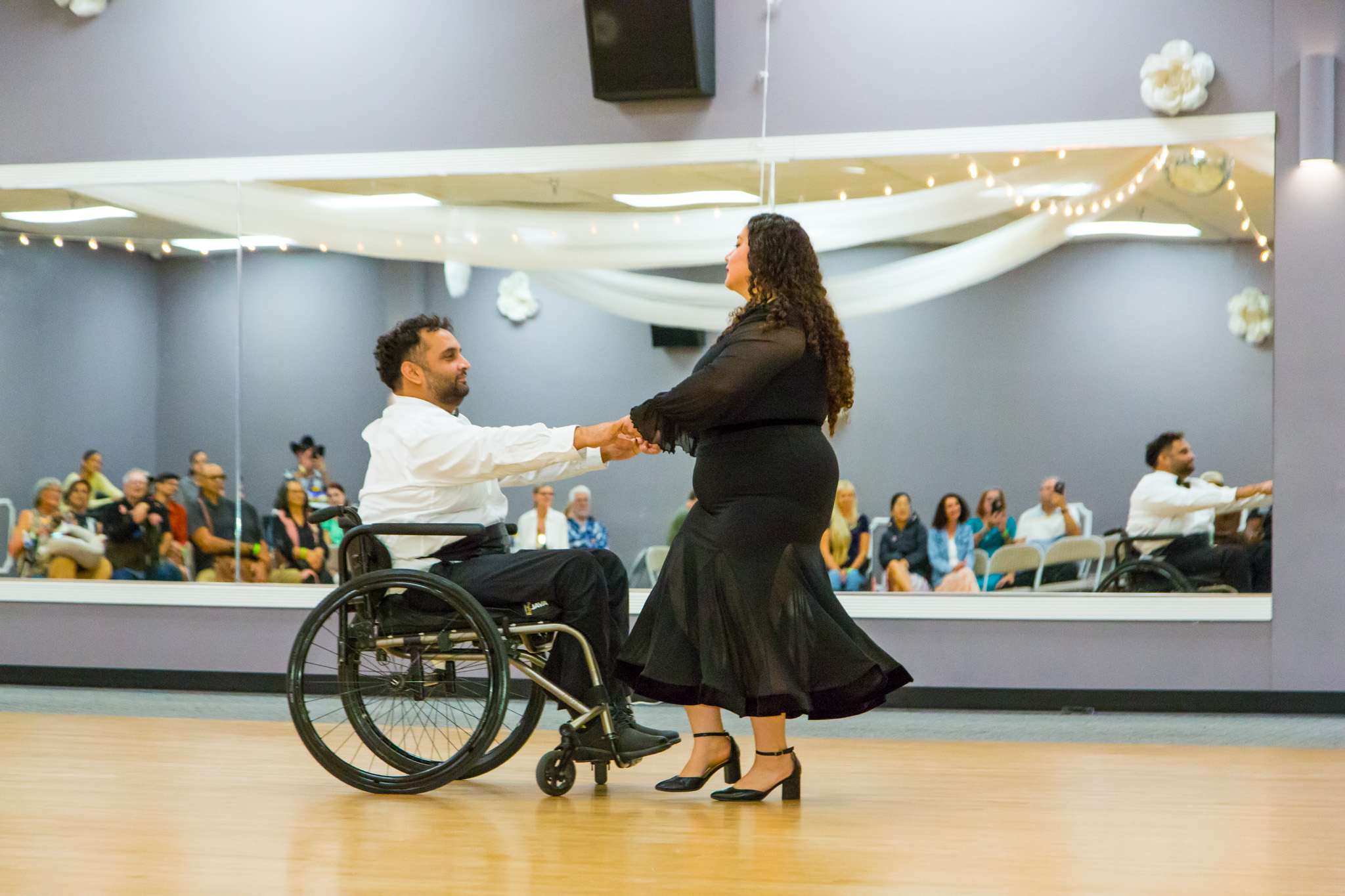 A man in a wheelchair and a woman dance together in a large room with an audience, mirror, and decorations.