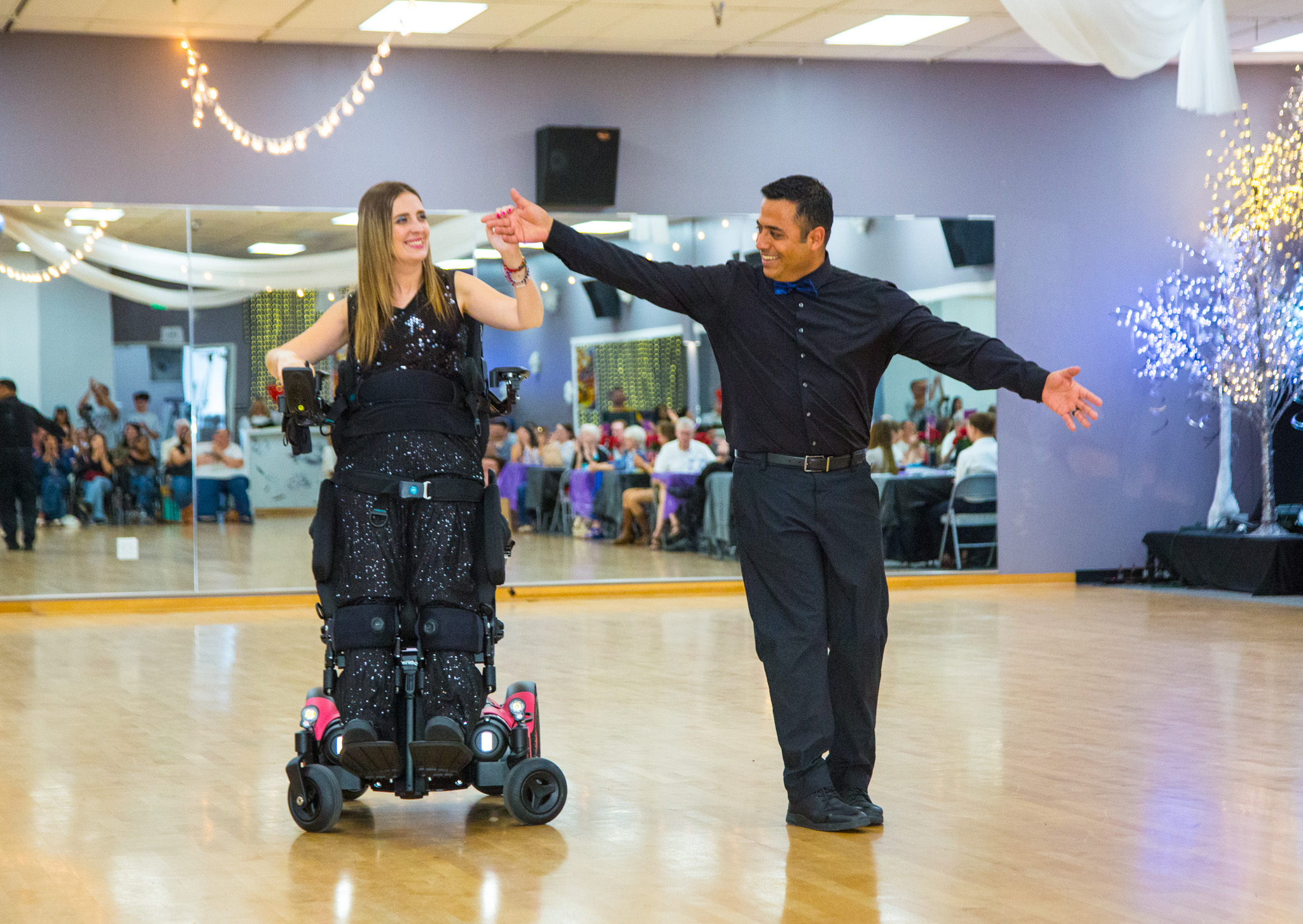 Person in a wheelchair dancing with a man in a dance hall, with guests and decorations in the background.