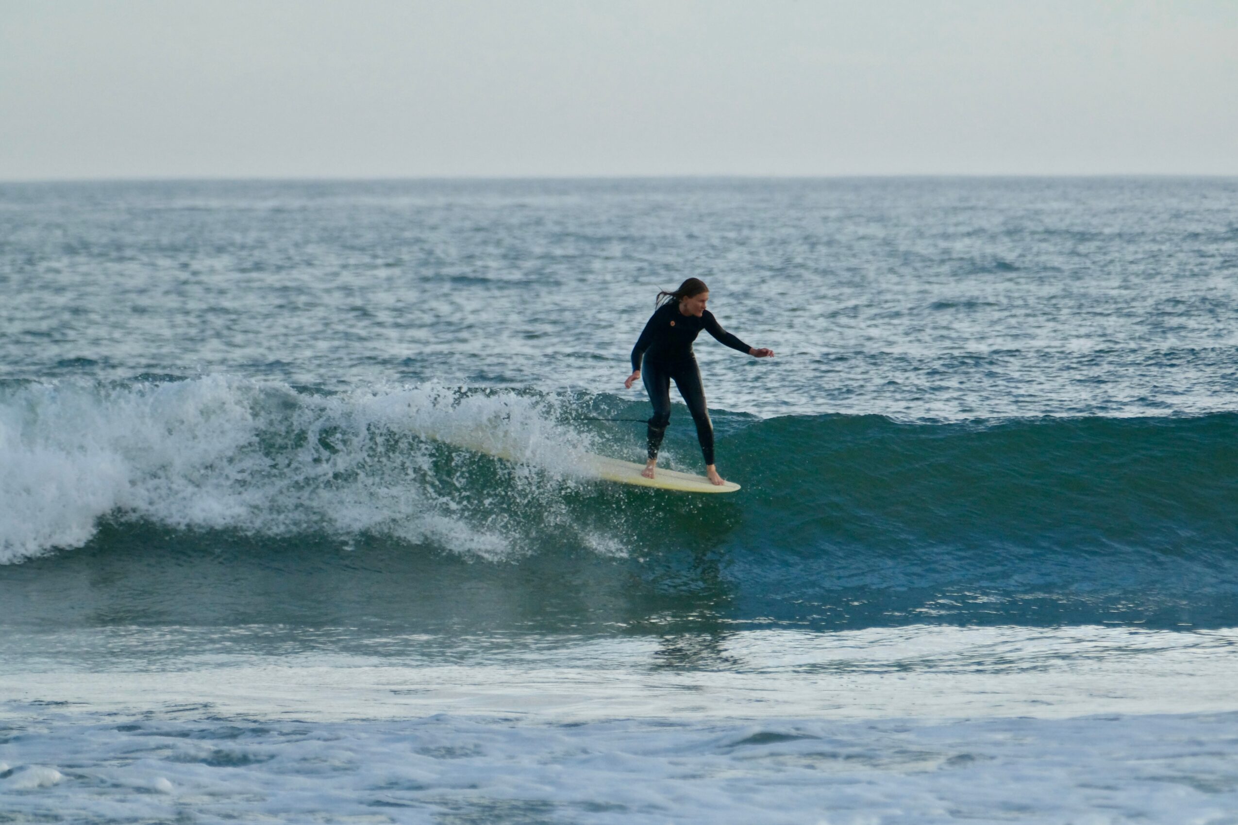 Person surfing on a wave in the ocean, wearing dark clothing, with a cloudy sky background.