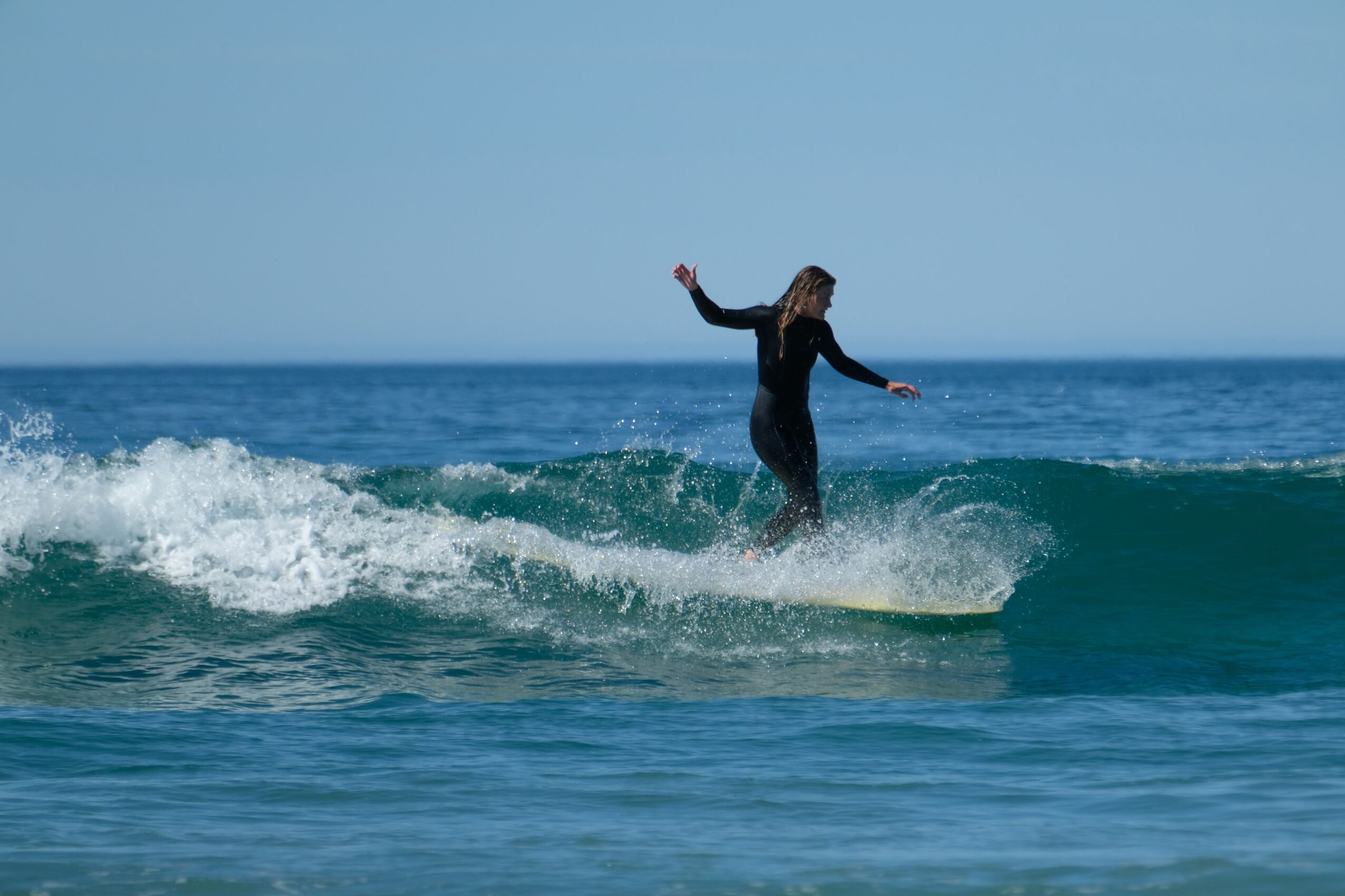 Person surfing on a wave in the ocean, with arms outstretched, against a clear sky.