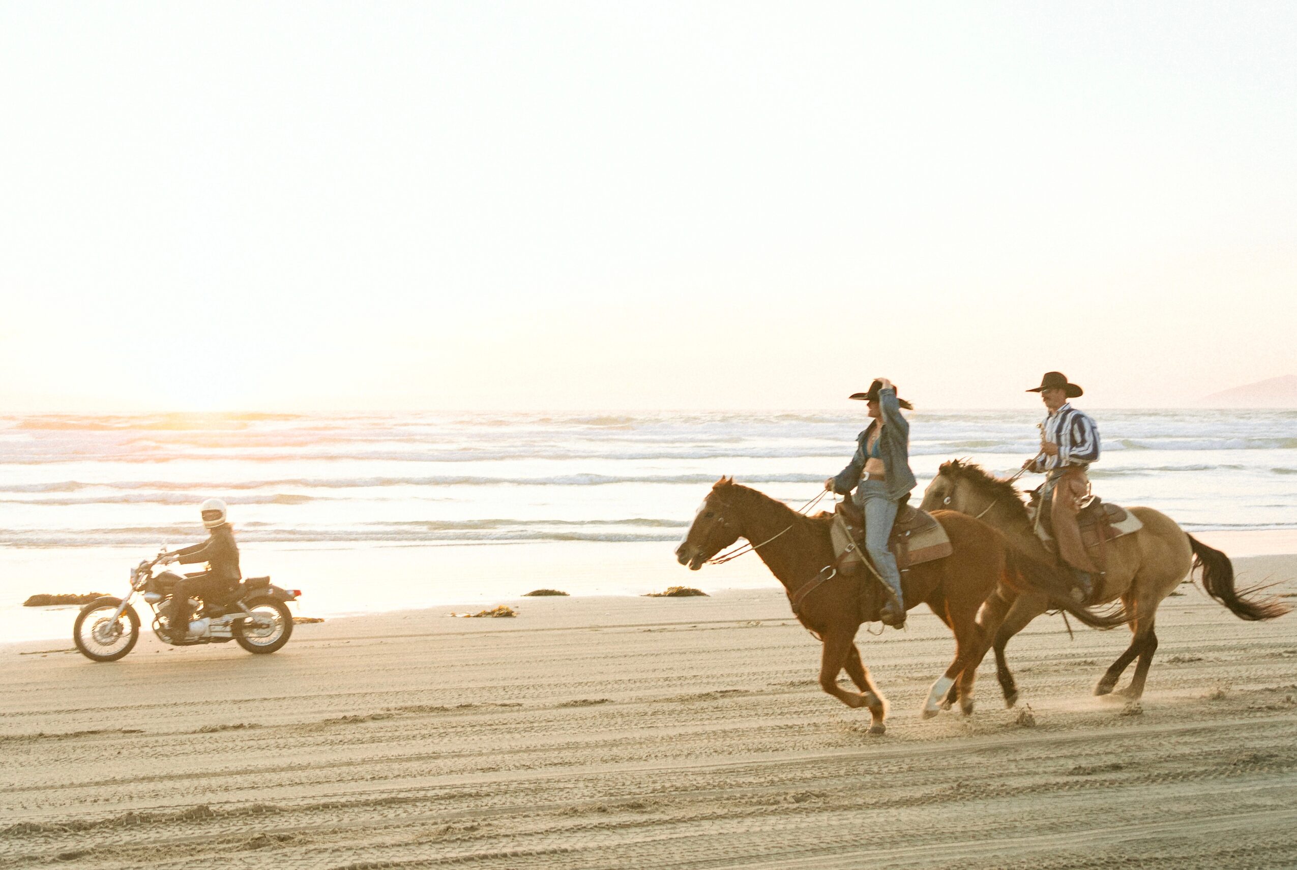Two people riding horses along the beach with a person on a motorcycle nearby, ocean waves in the background.