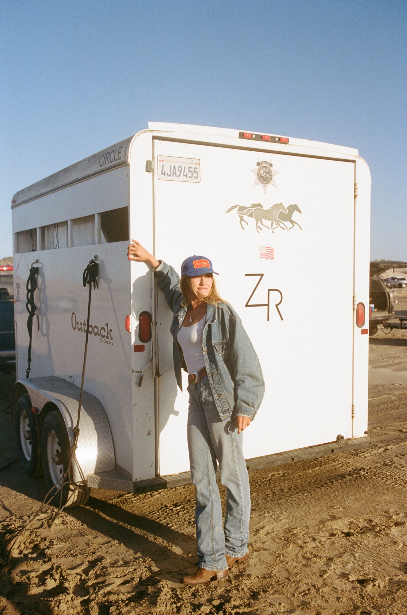 Person standing next to a white horse trailer, wearing a cap and casual clothing, on a dirt surface under a clear sky.