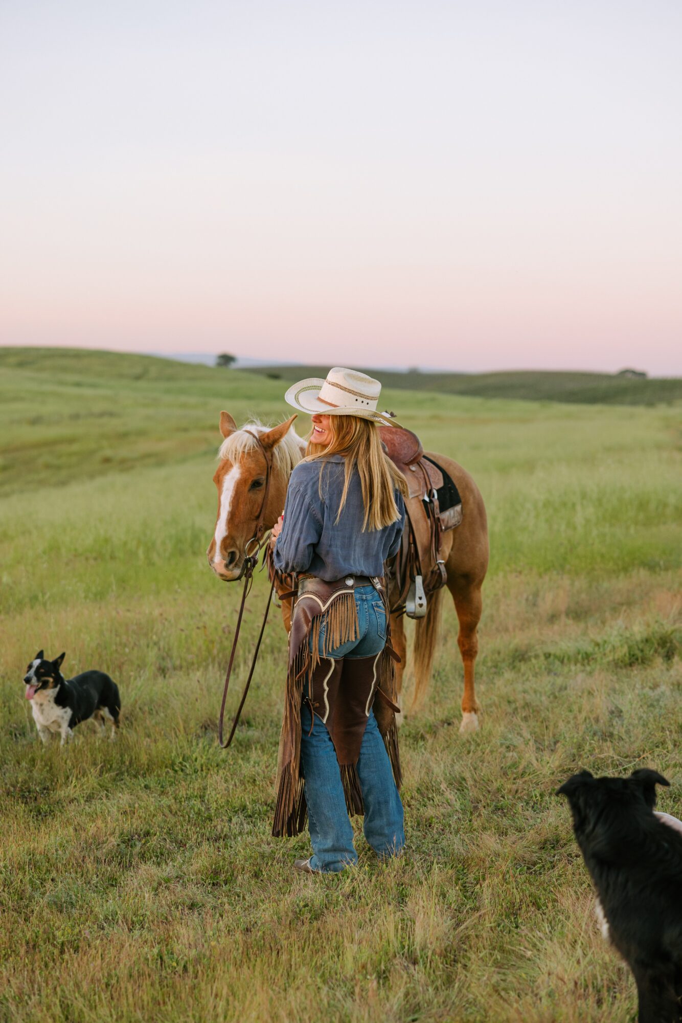 Person with a hat and fringed jacket stands in a grassy field with a horse, a dog, and a black animal nearby.