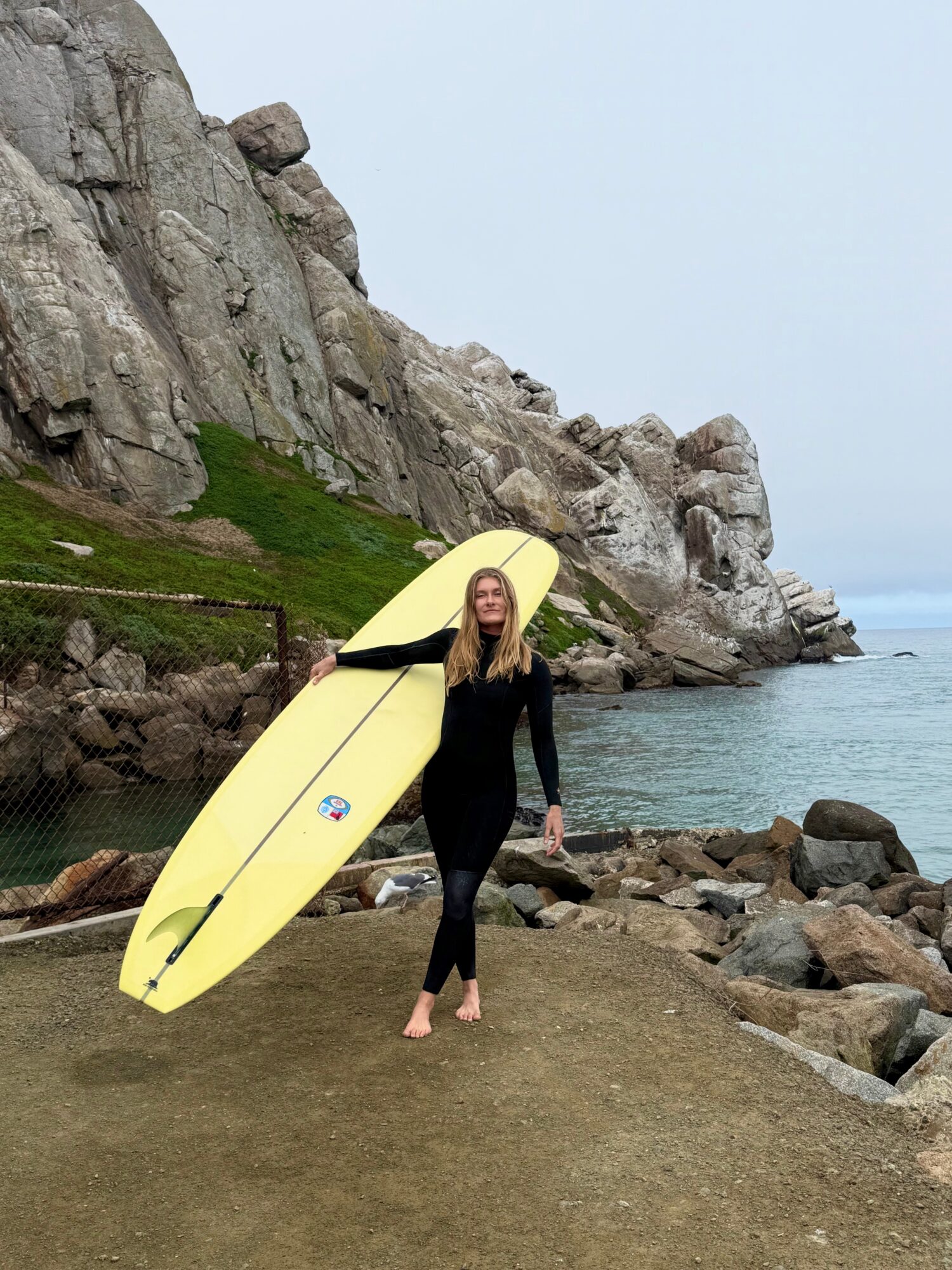 Woman standing on rocky shoreline holding a yellow surfboard, with cliffs and water in the background.