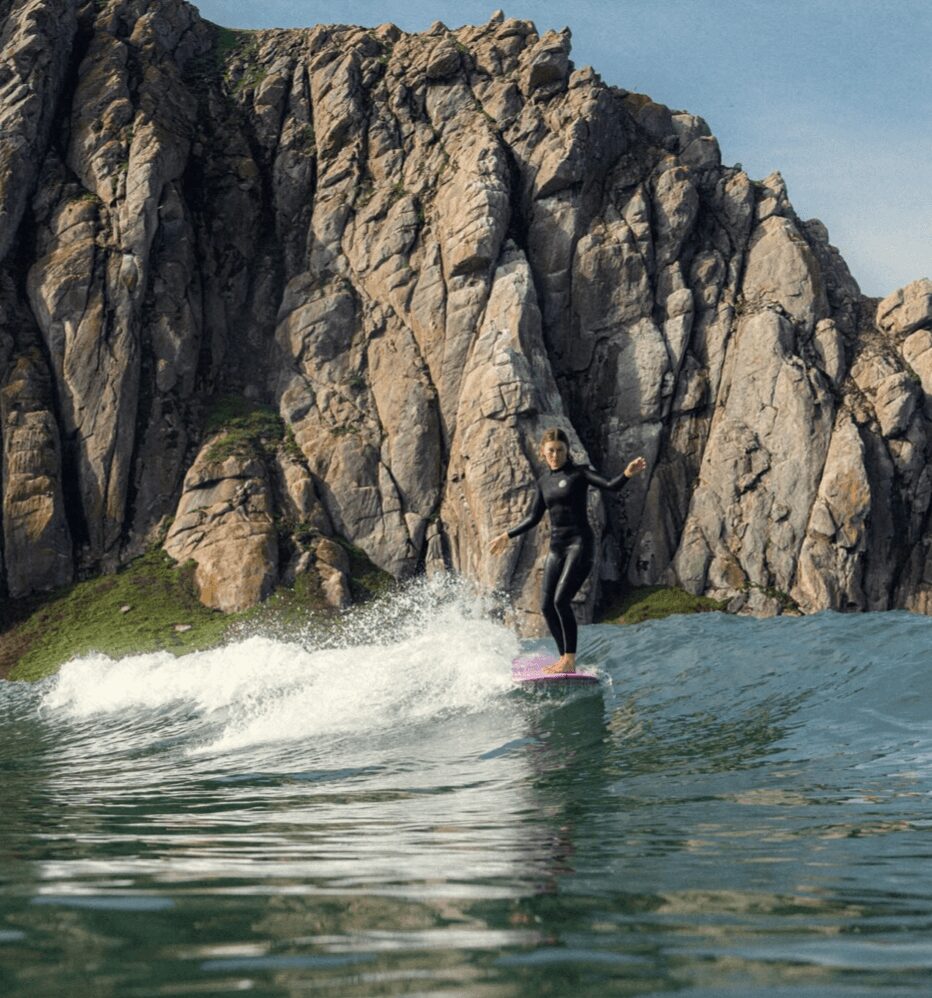 Person surfing on a wave near rocky cliffs, wearing a wetsuit, with water splashing around.
