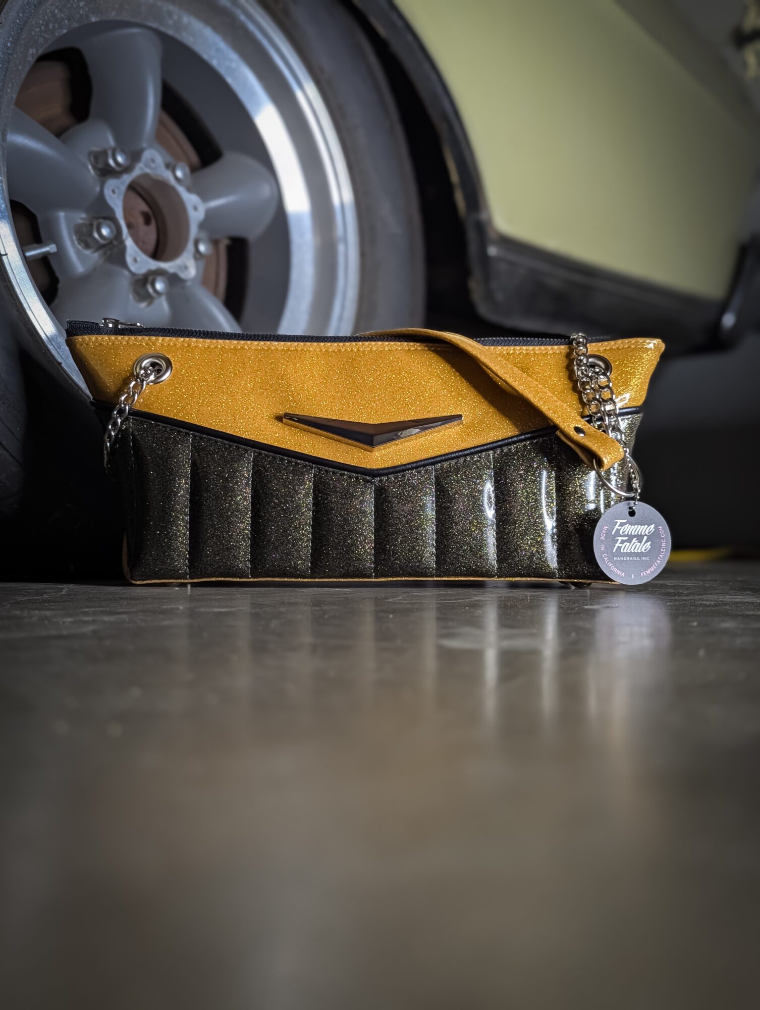 Yellow and black clutch purse with a chain strap and a round tag, placed on a floor near a car wheel.
