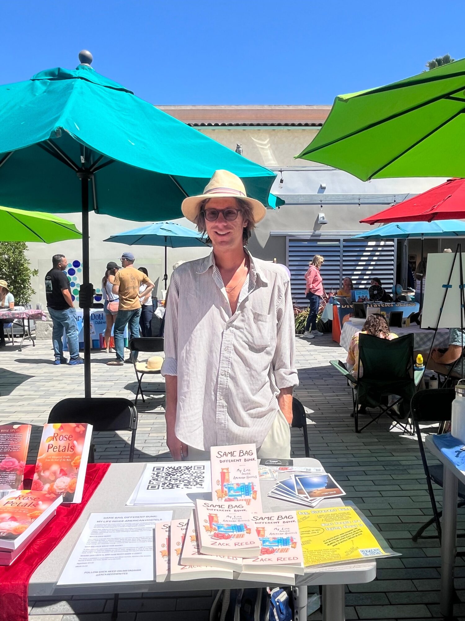 Person standing at outdoor table with pamphlets and papers, under colorful umbrellas, with city buildings in background.