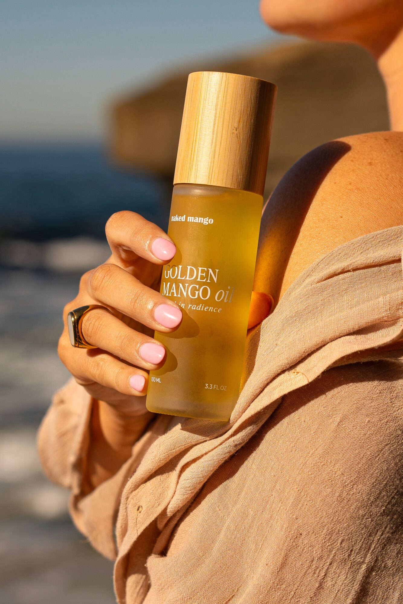 Person holding a yellow bottle of Golden Mango oil with a wooden cap at the beach, ocean in background, sunlight on skin.