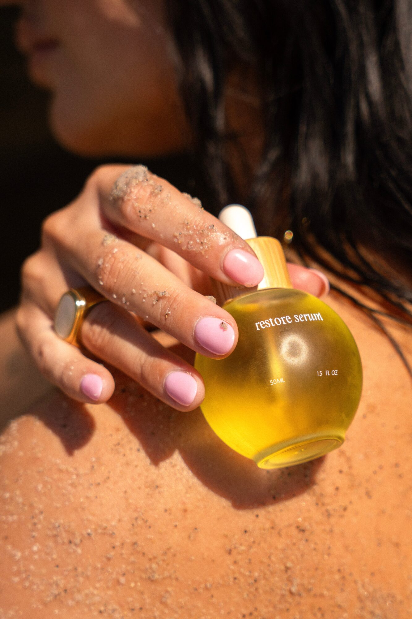 Person holding a small yellow bottle labeled 'restore serum' with fingers covered in sand, close-up of hand and bottle.