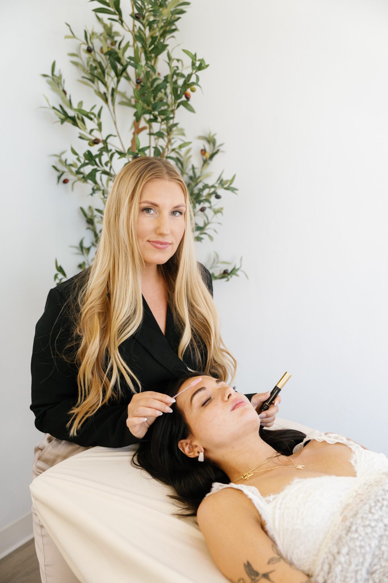 A woman with long blonde hair stands beside a woman lying on a treatment bed, receiving a facial treatment, in a bright room with a plant in the background.