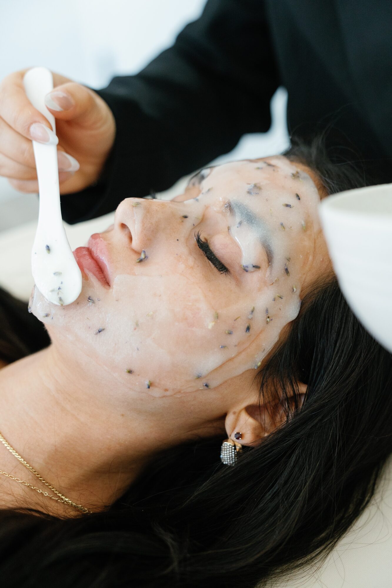 Person receiving facial treatment with a mask applied, lying down with eyes closed, a hand applying product with a spatula, and a white bowl nearby.