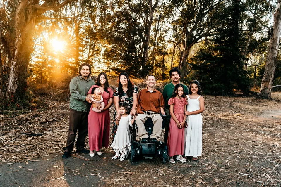 Group of seven people standing outdoors in a park during sunset, with trees in the background, smiling and posing for the photo.