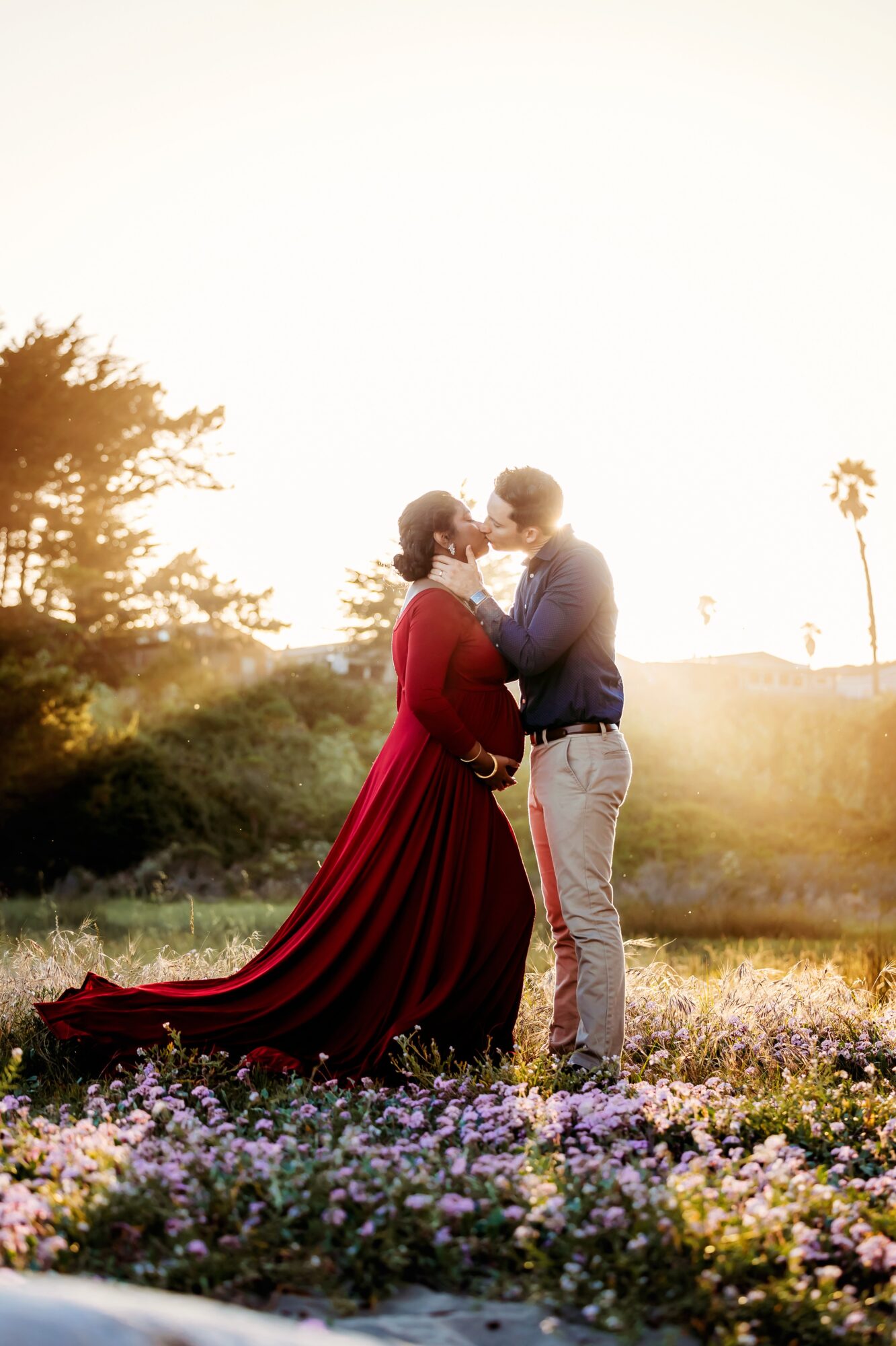 A couple stands close together outdoors during sunset, holding hands and looking at each other, with flowers in the foreground.