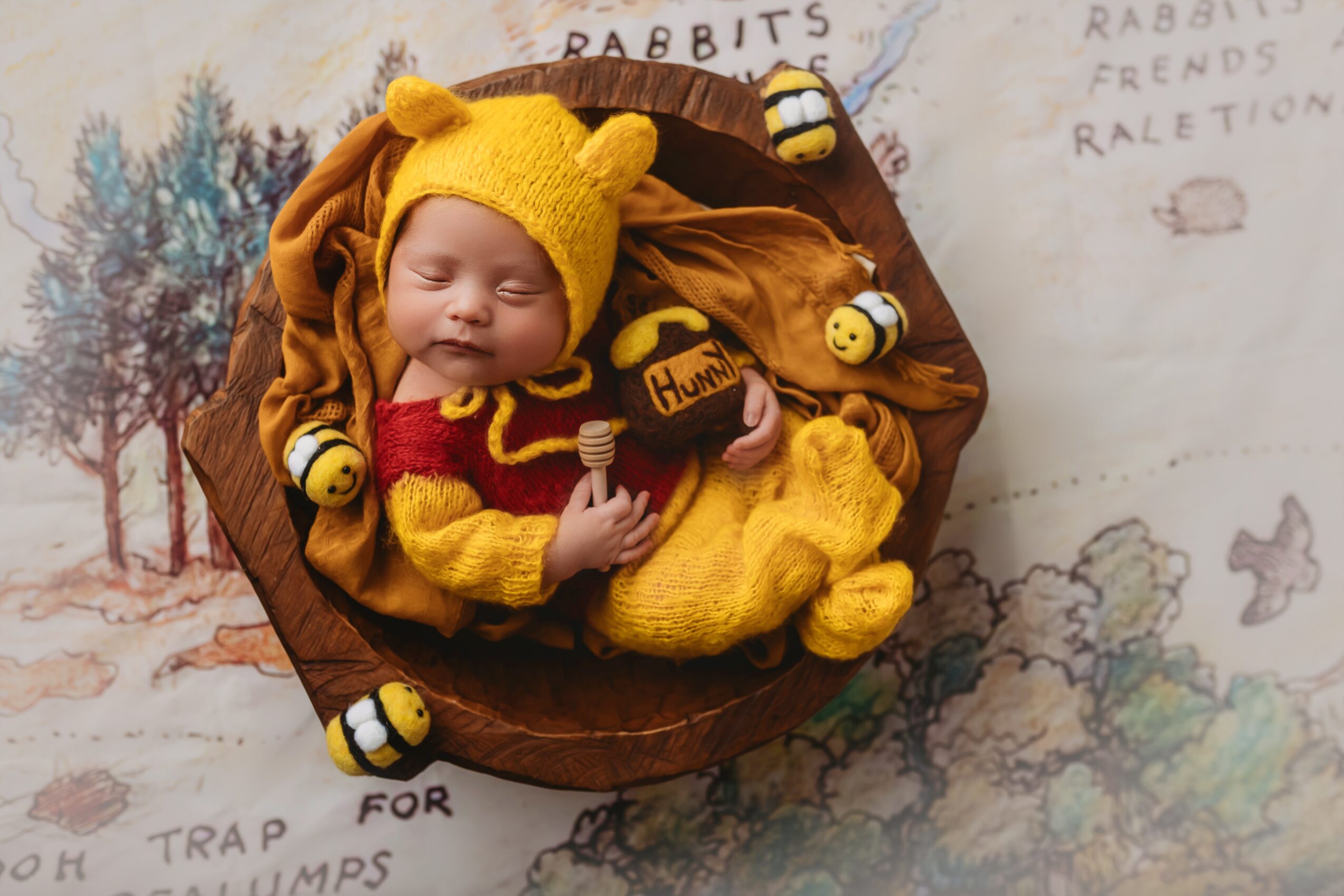 Baby dressed in yellow bear costume with red shirt, lying in a brown basket, surrounded by small bee plush toys.