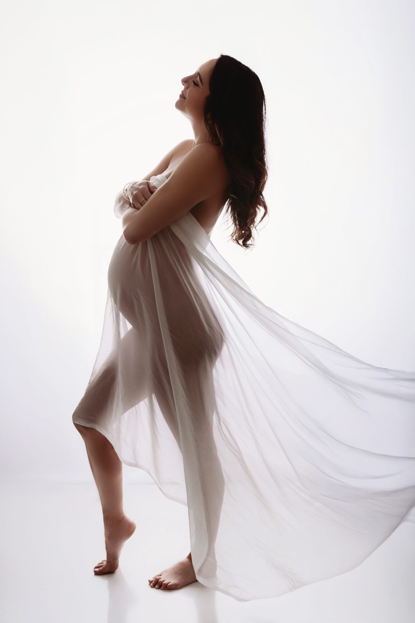 Pregnant woman standing sideways, draped in sheer fabric, with arms crossed and head tilted upward.