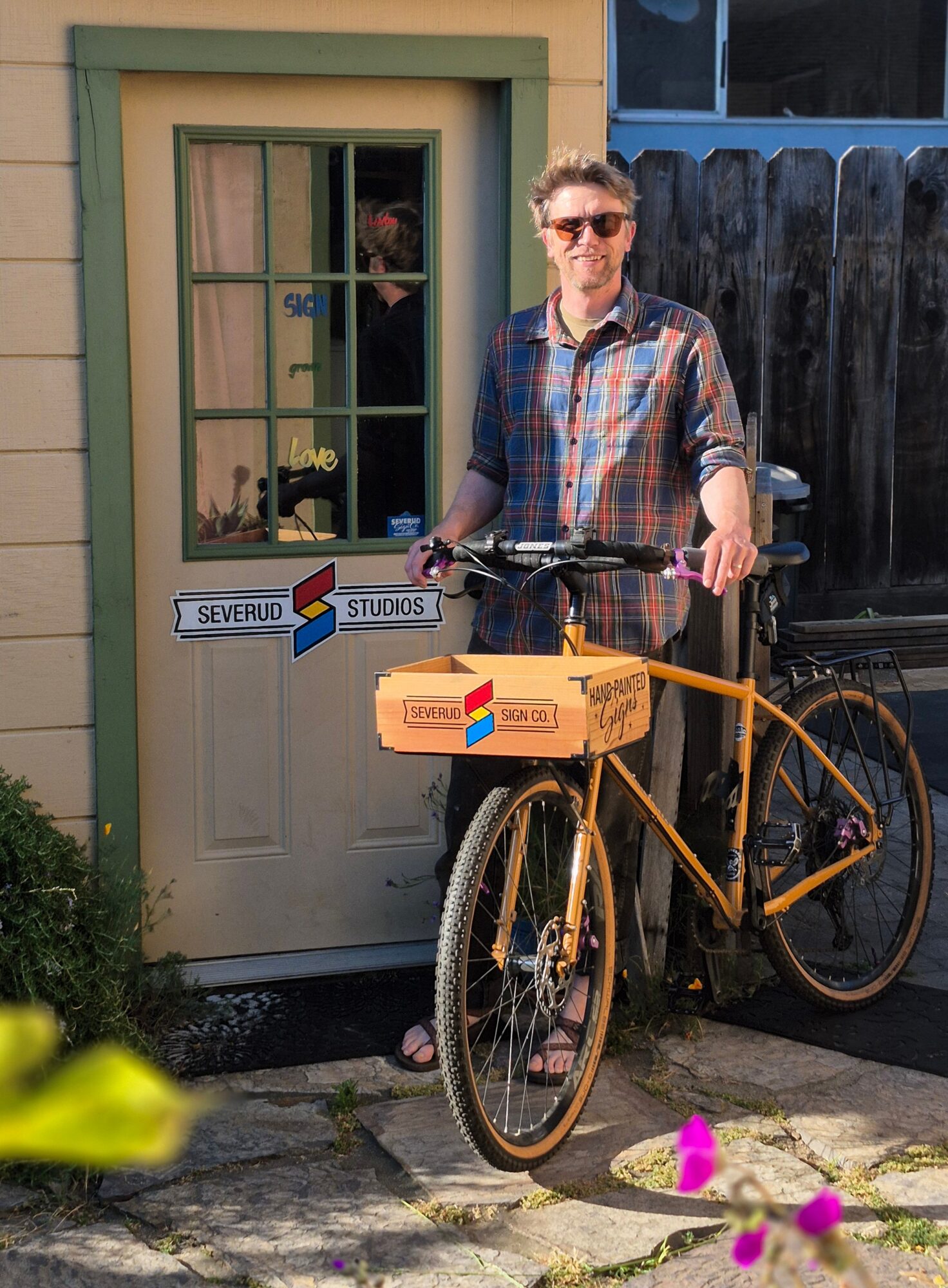 Man with sunglasses stands next to a yellow bicycle with a cardboard box, in front of a door with a sign.