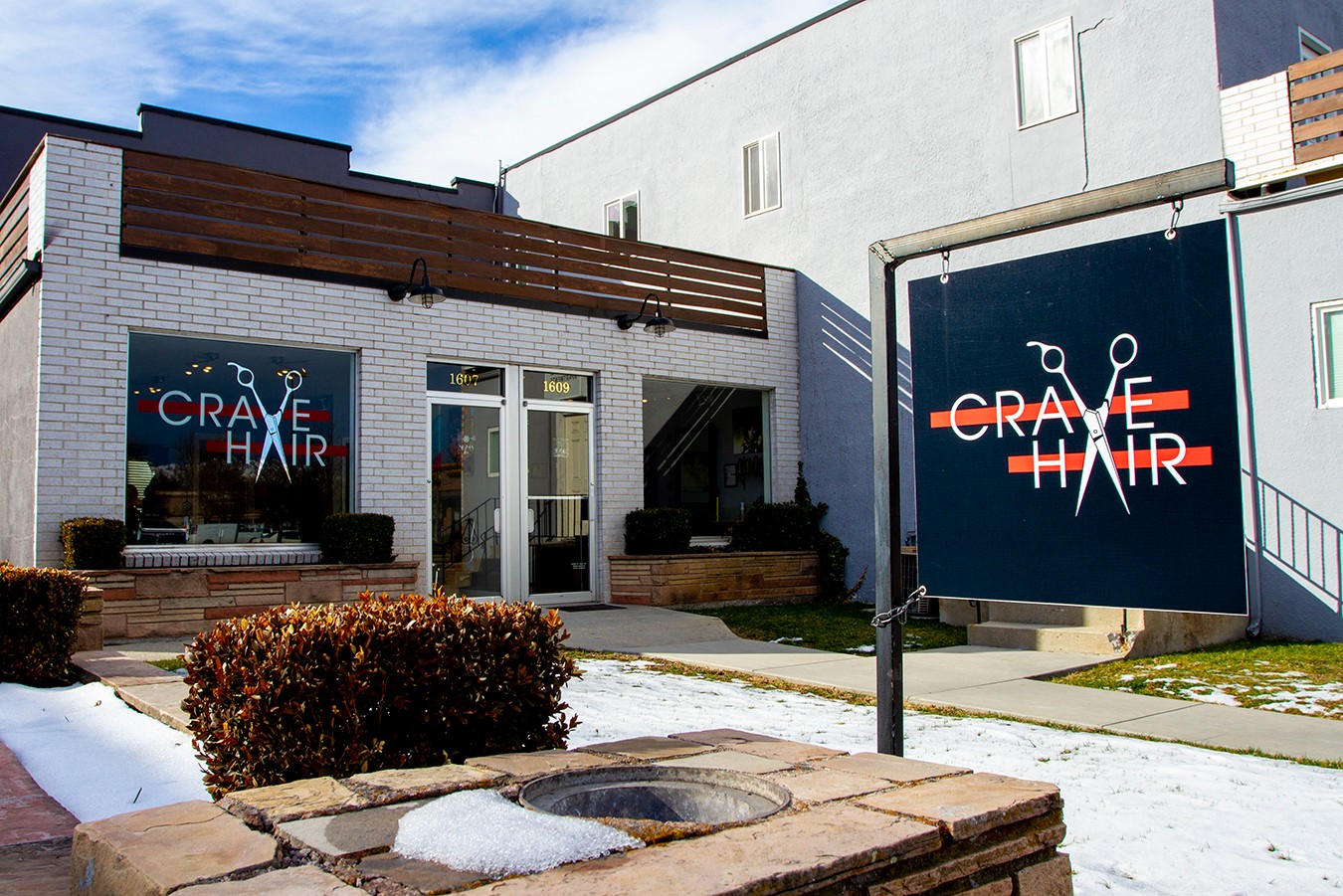 Exterior of a building with a sign and window displaying the word 'CRAVE HAIR' with a red and blue background, bushes, and a sidewalk.