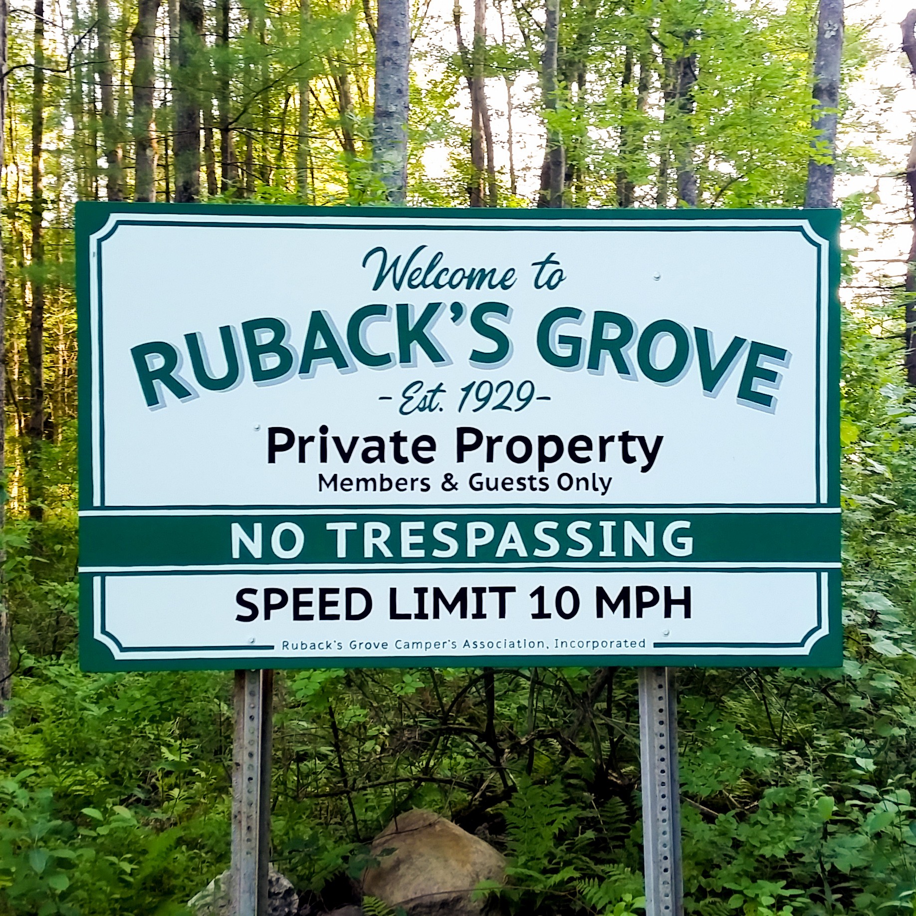 Sign in a forested area with trees and greenery, welcoming visitors to Ruback's Grove, indicating private property, no trespassing, and a 10 MPH speed limit.