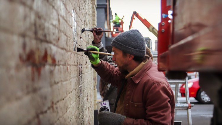 Two workers repair a brick wall outdoors, one using a tool, with construction equipment in the background.