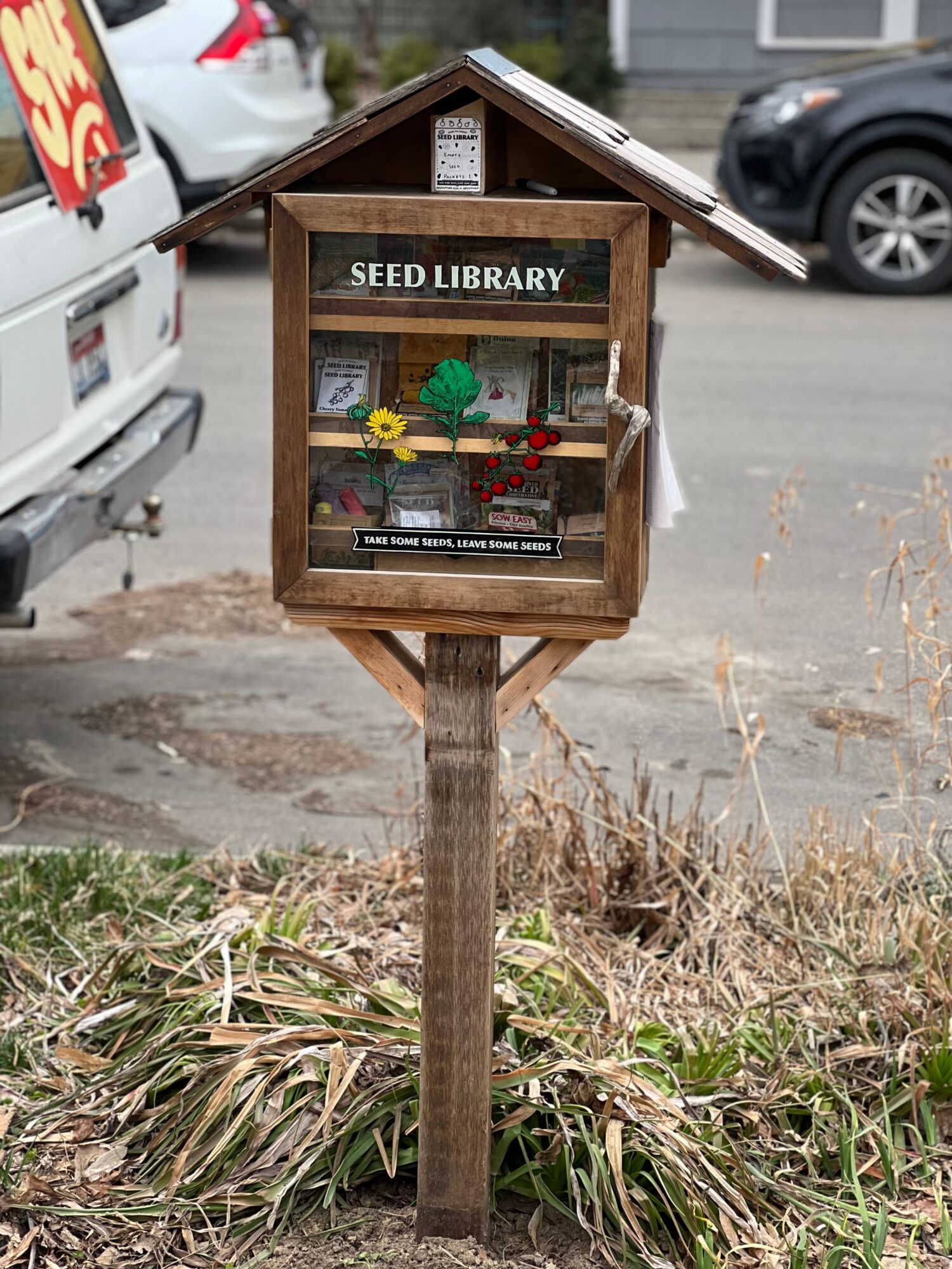 Small wooden seed library box on a post with books and decorations inside, located outdoors near parked cars.