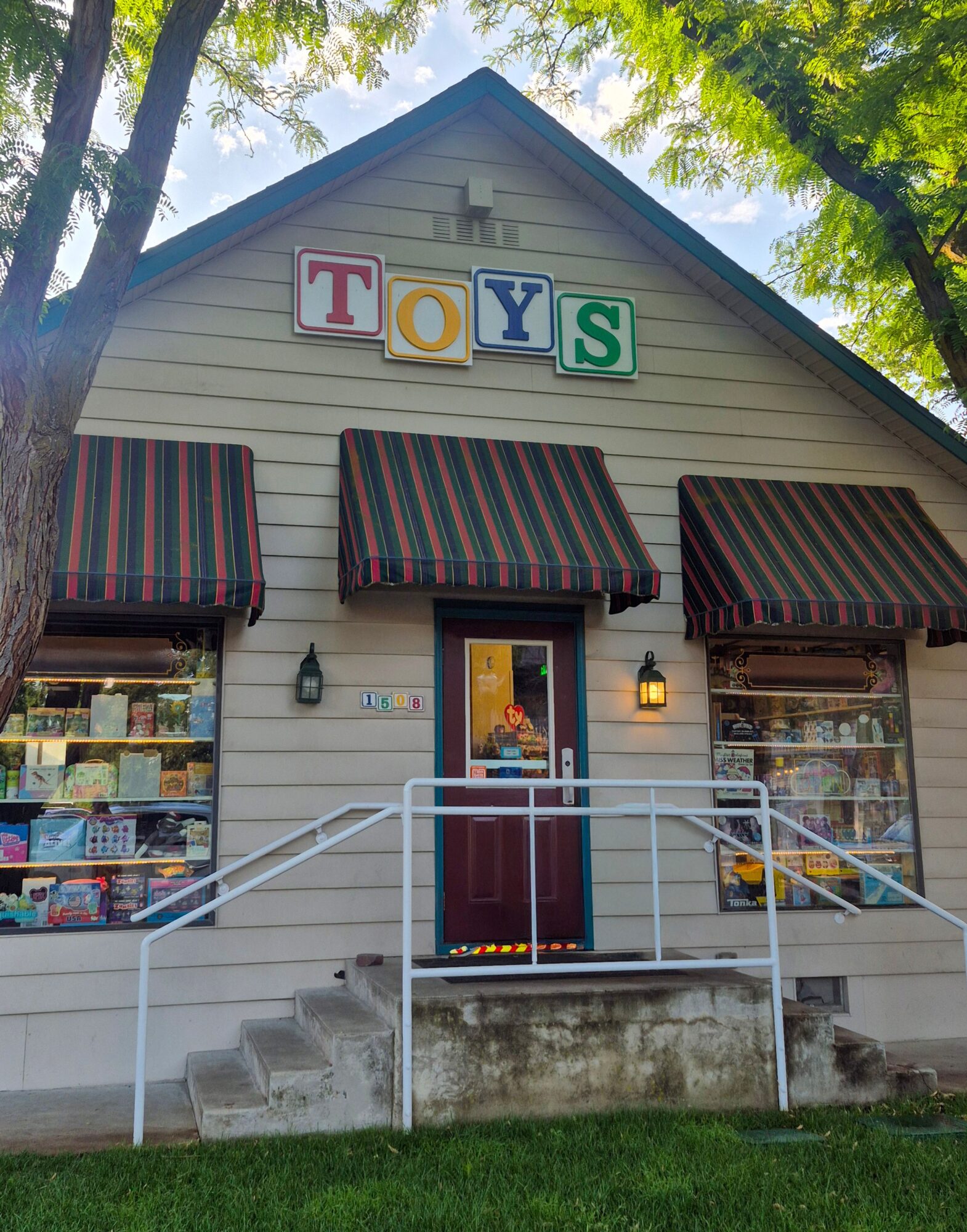 Small toy store with striped awnings, colorful sign, and glass windows, surrounded by trees and greenery.