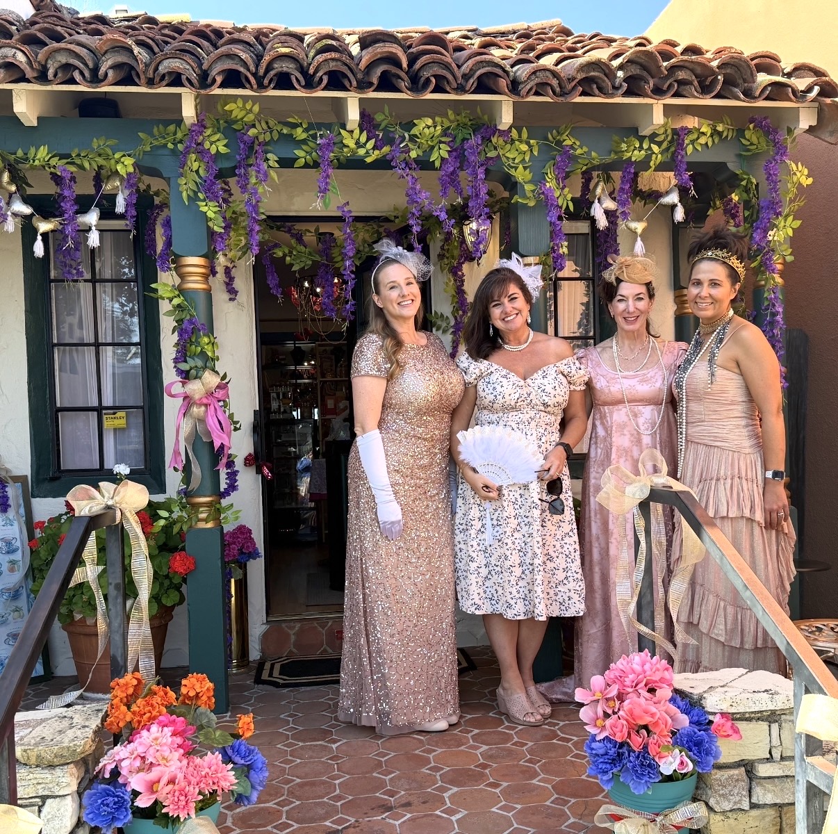 Four women in elegant dresses stand under decorated archway with flowers and greenery, smiling outside a building.