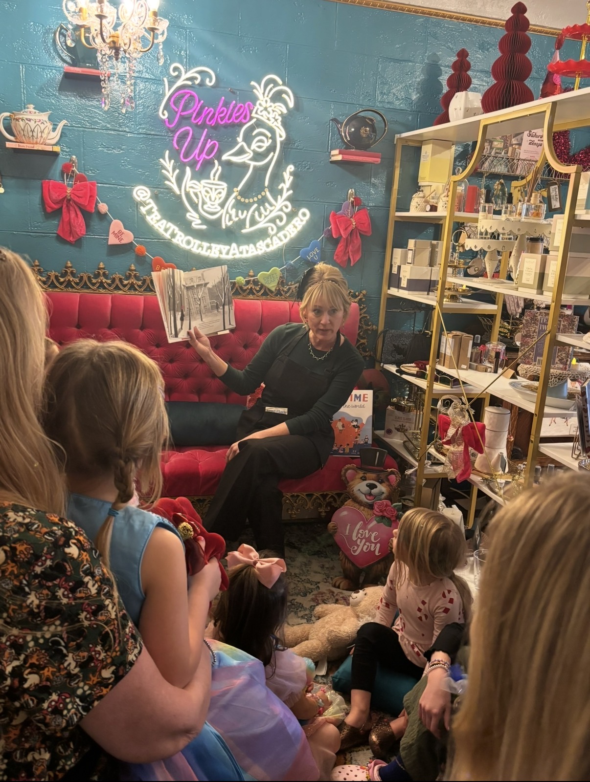 A woman reading a book to children in a decorated room with pink bows, a neon sign, and shelves filled with toys and gifts.