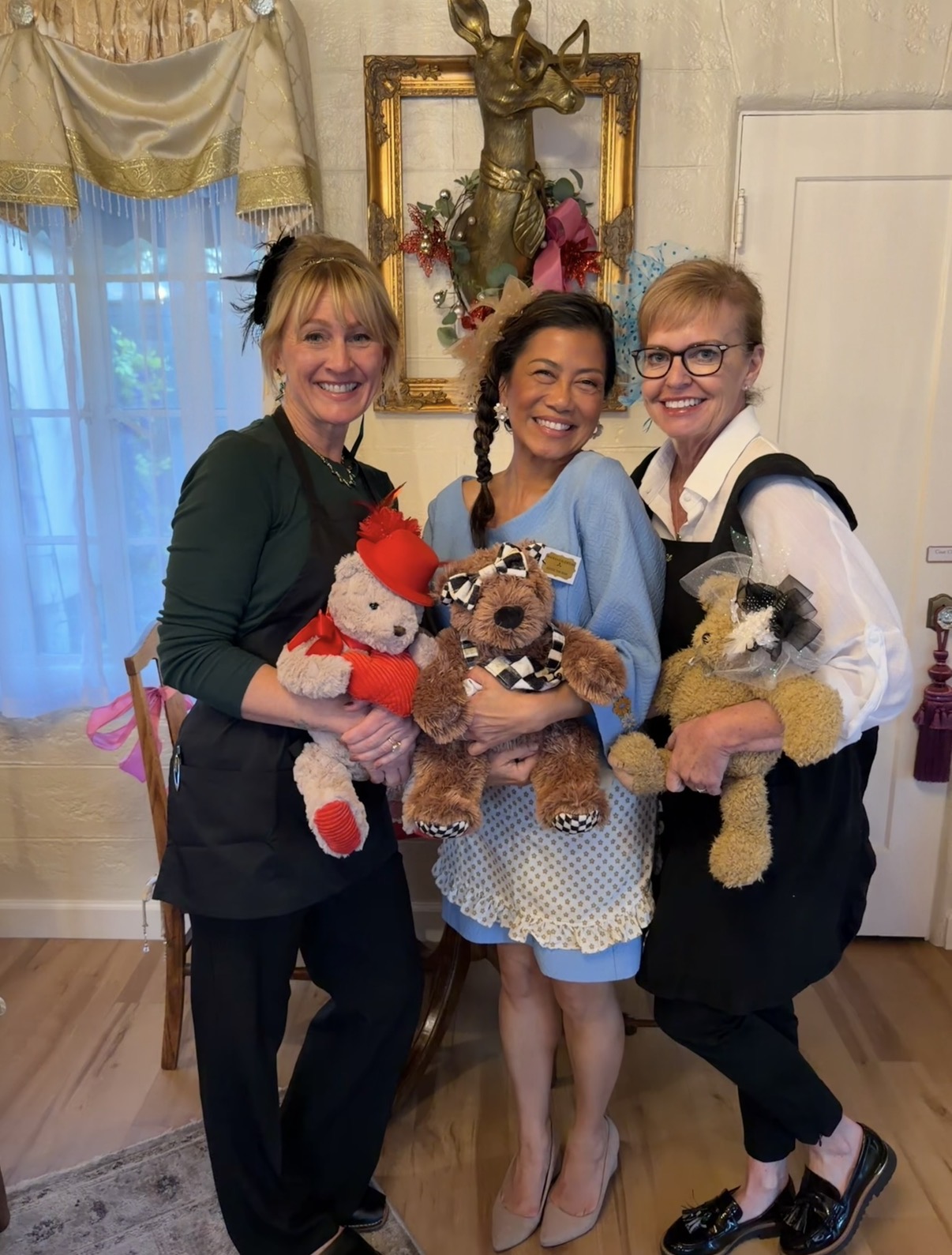 Three women smiling indoors, holding stuffed animals, standing in front of a decorative wall with a framed artwork and a window.