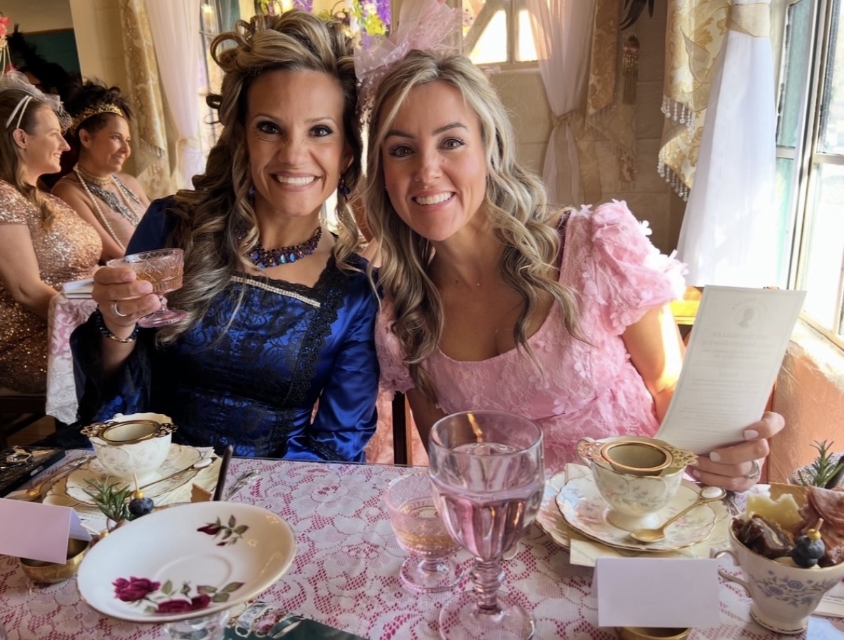 Two women smiling at a table with teacups, plates, and a glass, in a decorated room with sunlight.