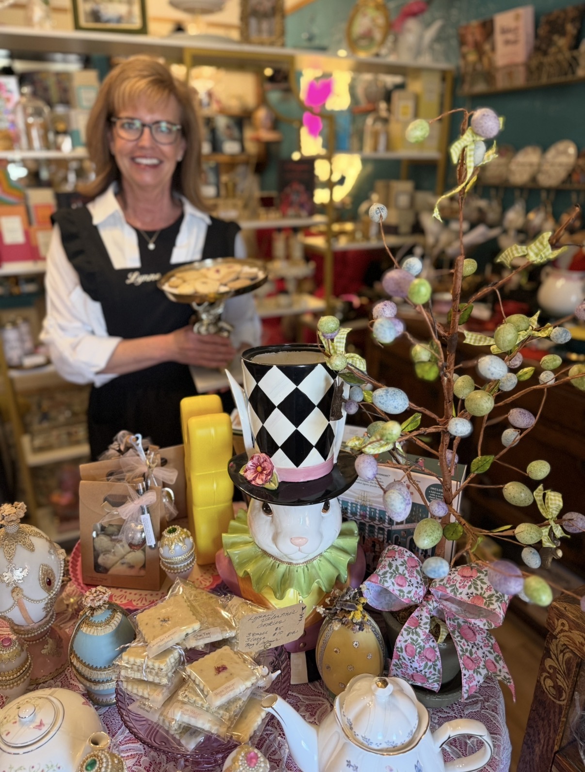 Woman with glasses and a black apron stands behind a table with decorated eggs and a tall branch with colorful eggs and ribbons.