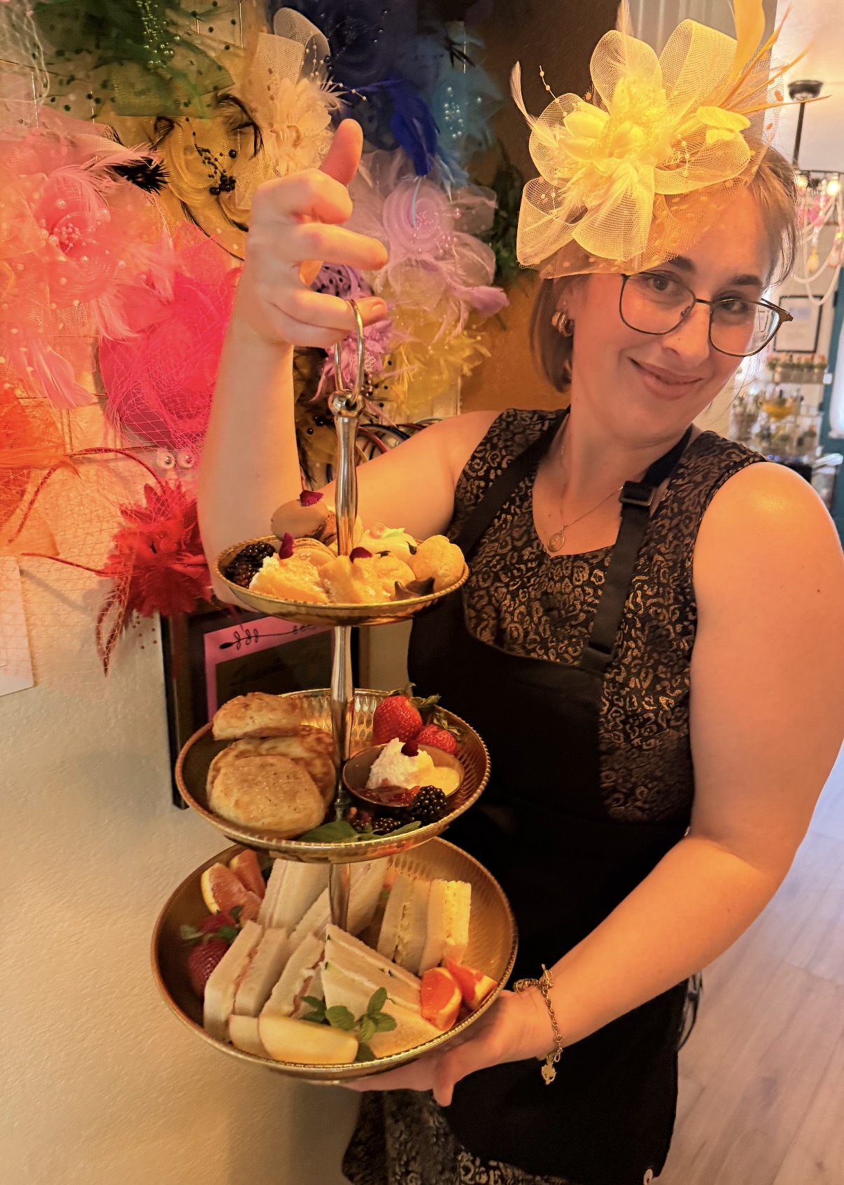 Woman with glasses and large yellow flower headpiece holding a three-tiered tray of desserts, smiling indoors.