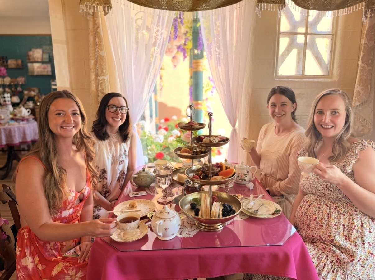 Five women sit around a table with food, smiling indoors with a window and curtains in the background.