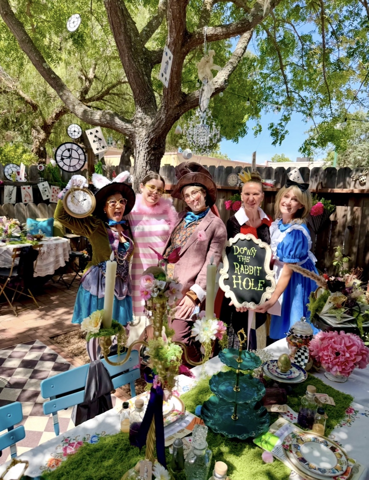 Group of children in costumes standing outdoors under a large tree with a fence and sky in background.