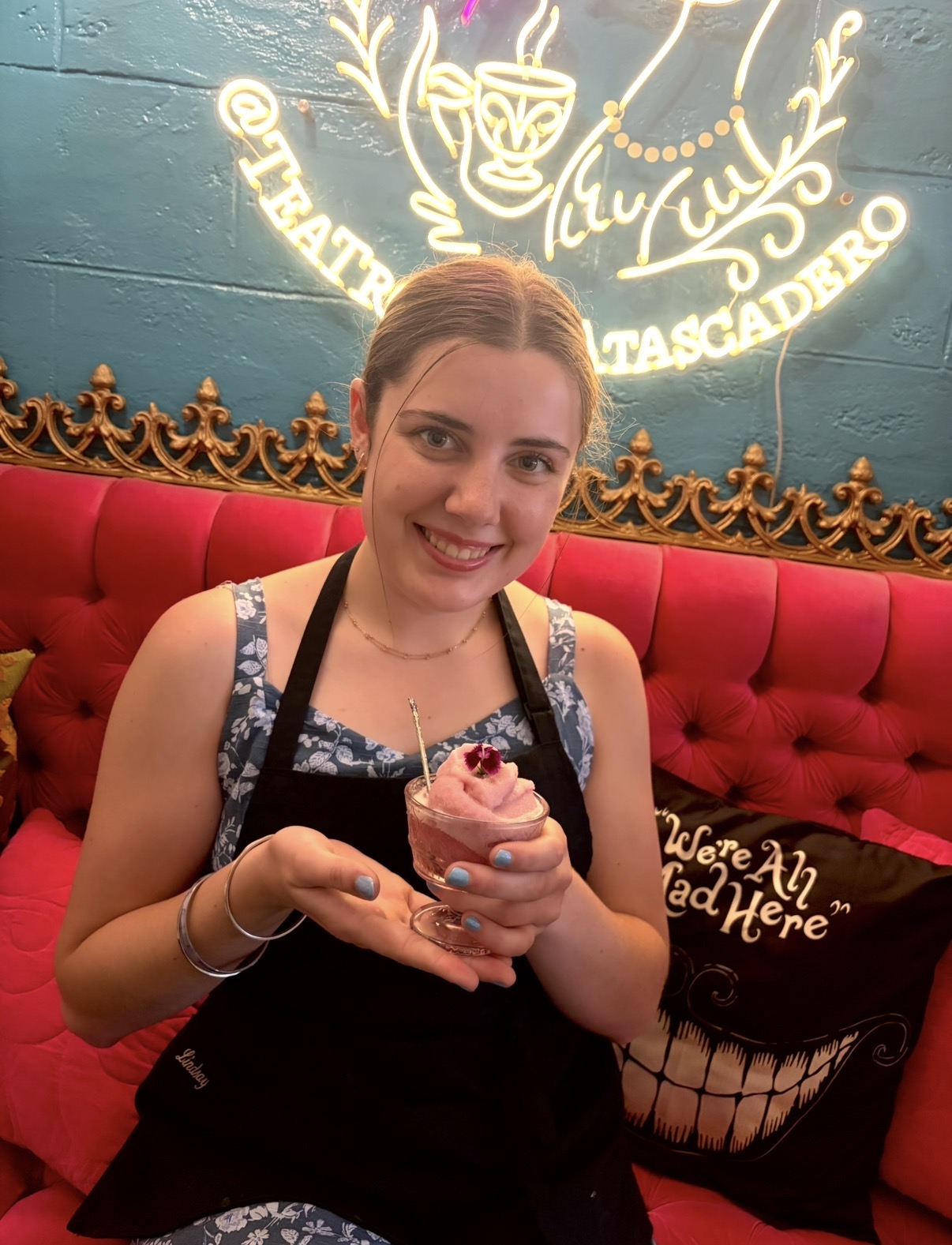 Young woman with a pink ice cream cone, smiling, sitting on a red cushioned bench, neon sign in background