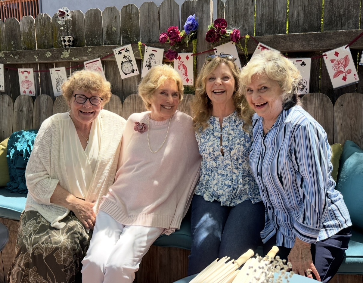 Four women sitting together outdoors, smiling, with birthday decorations and flowers in the background.