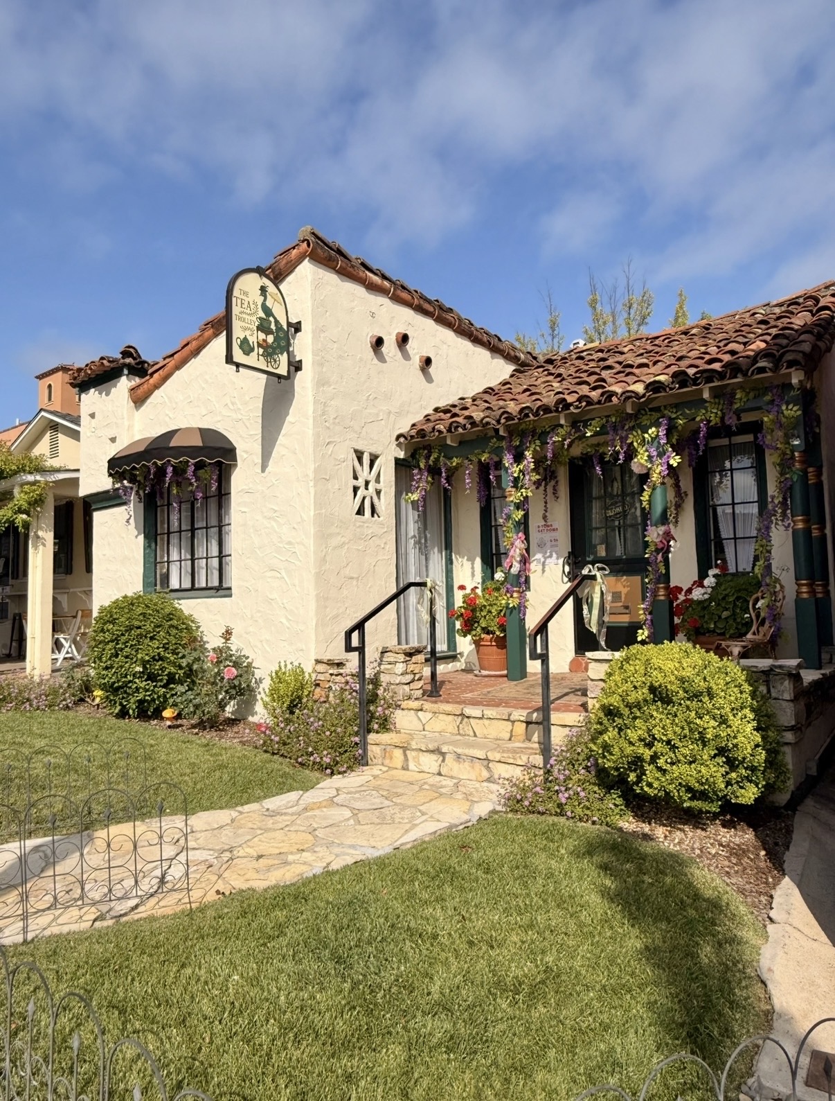 Two charming buildings with tiled roofs, surrounded by greenery and flowers, under a partly cloudy sky.