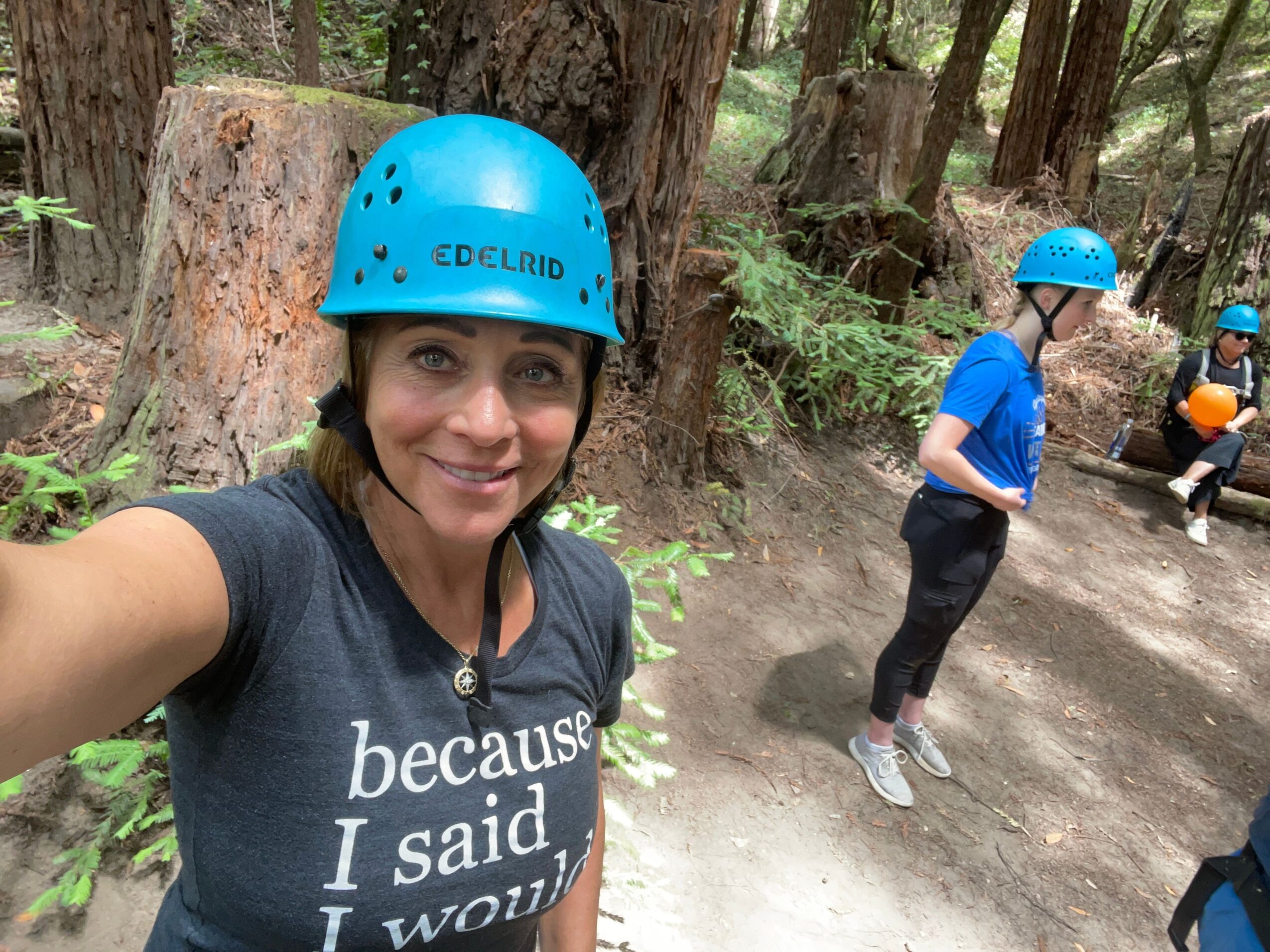 Person wearing a blue helmet and black shirt taking a selfie outdoors with a person climbing a rock wall in the background.