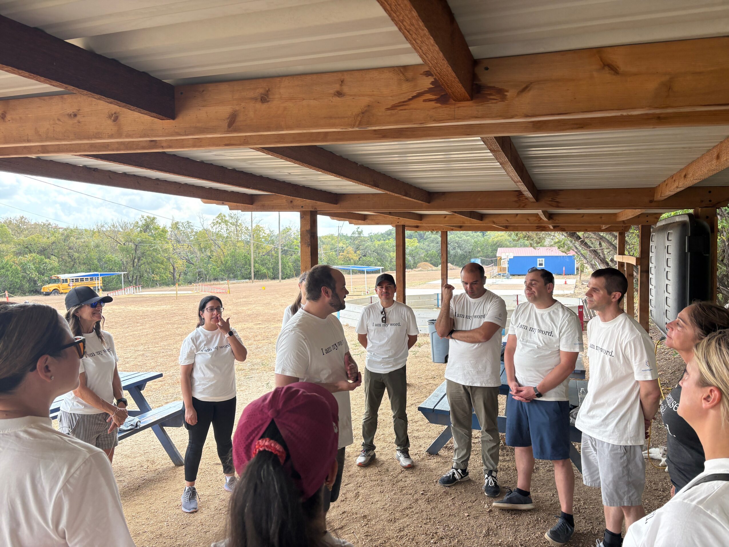 Group of people standing in a semi-circle outdoors under a wooden shelter, engaging in conversation.