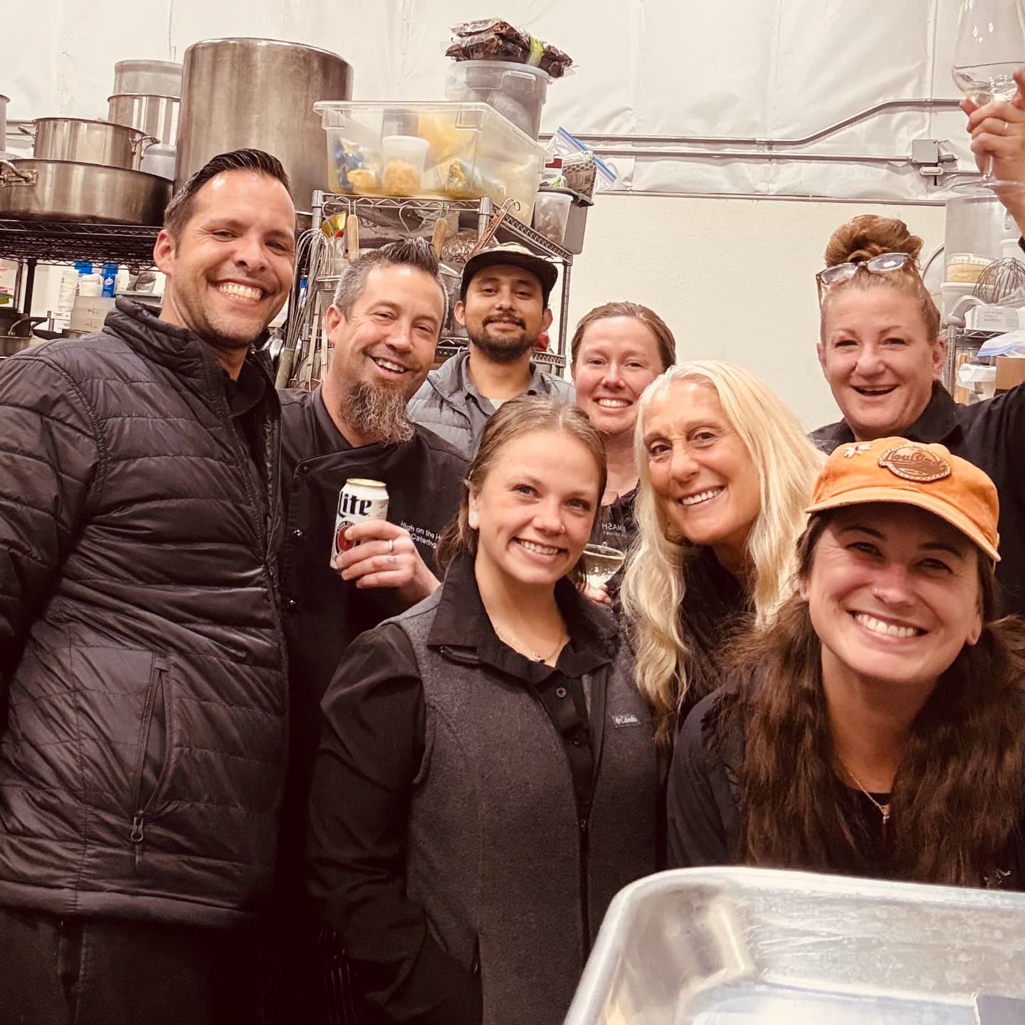 Group of nine smiling people in a kitchen, some holding drinks, with shelves and kitchen equipment in the background.