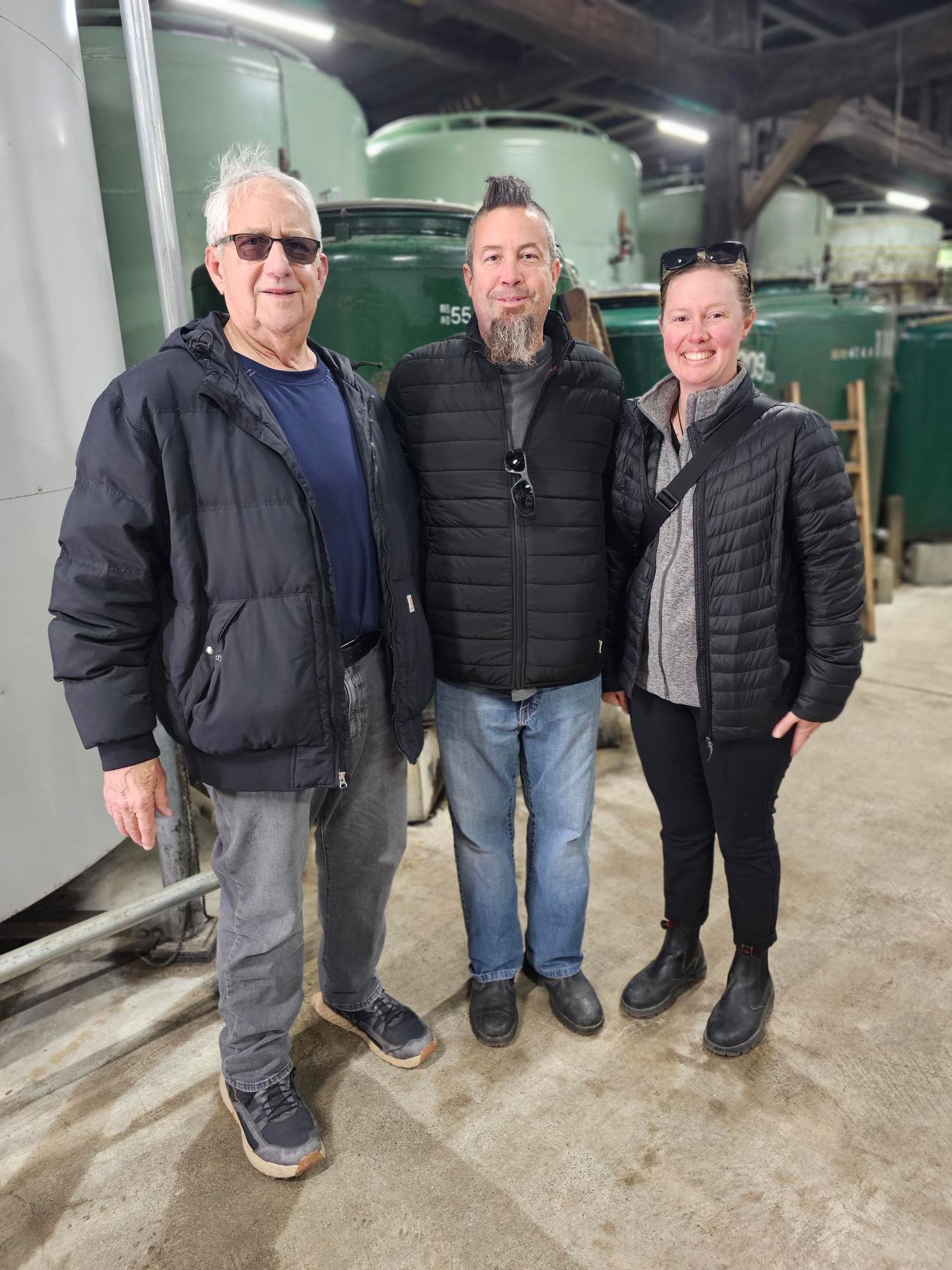 Three people standing indoors on a concrete floor, with industrial equipment in the background.