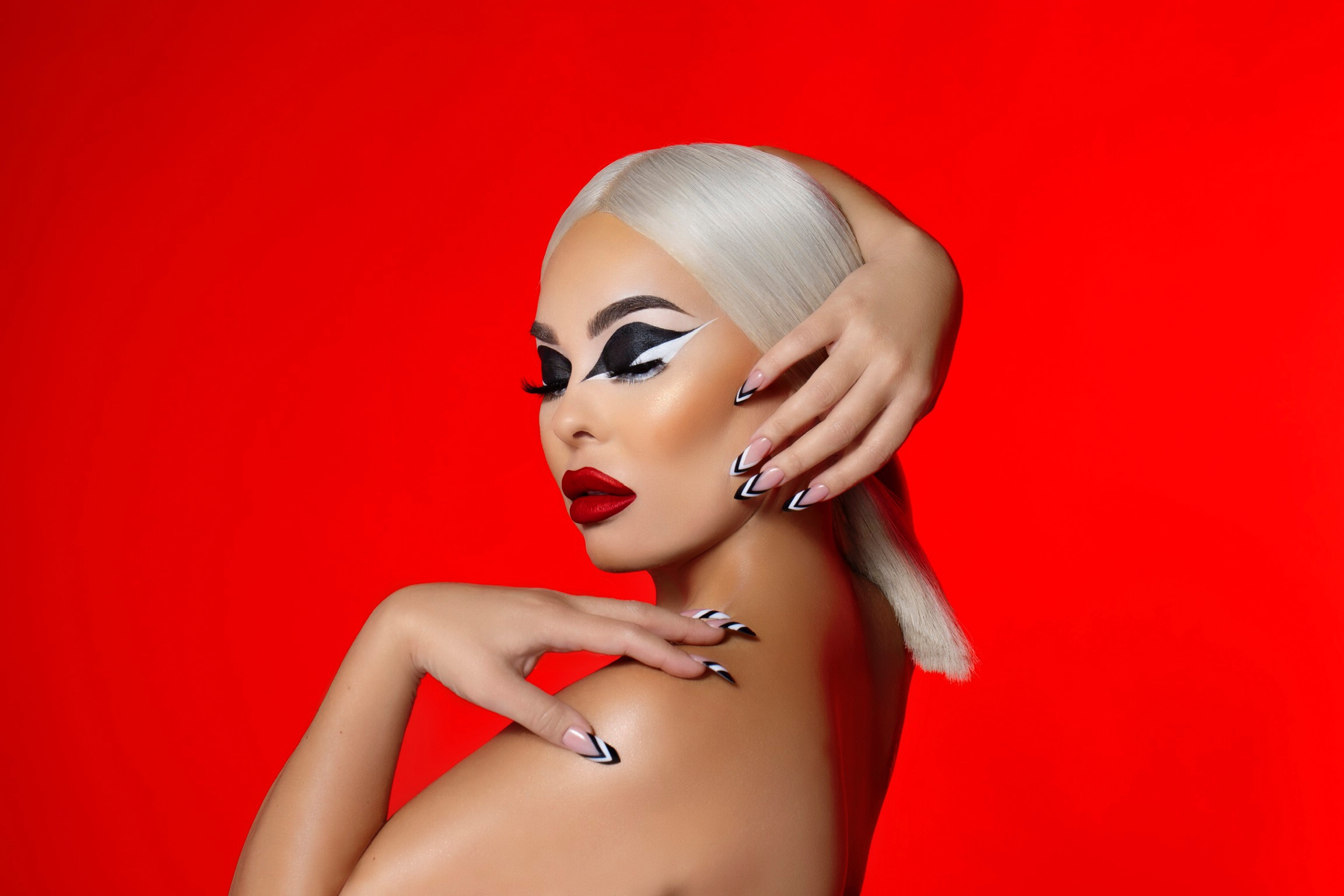 Woman with platinum blonde hair and bold makeup poses against a red background.