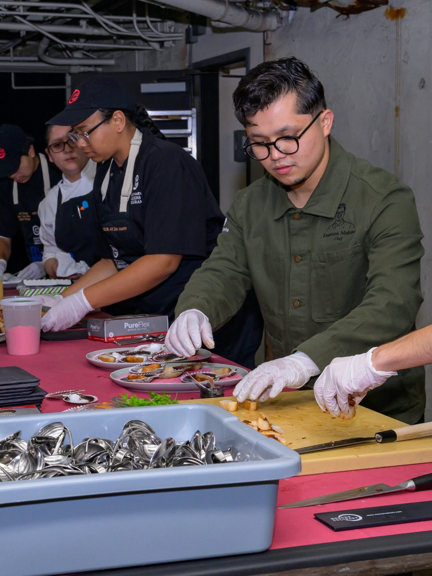People preparing food at a table with various utensils and ingredients, wearing gloves and aprons, in an indoor setting.