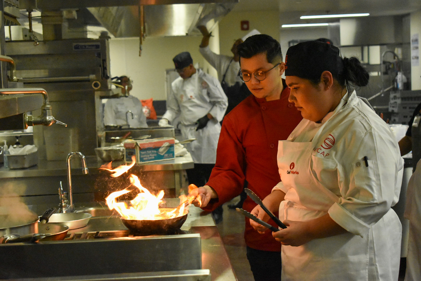 Two chefs in a kitchen cook with a flaming pan, while others work in the background.