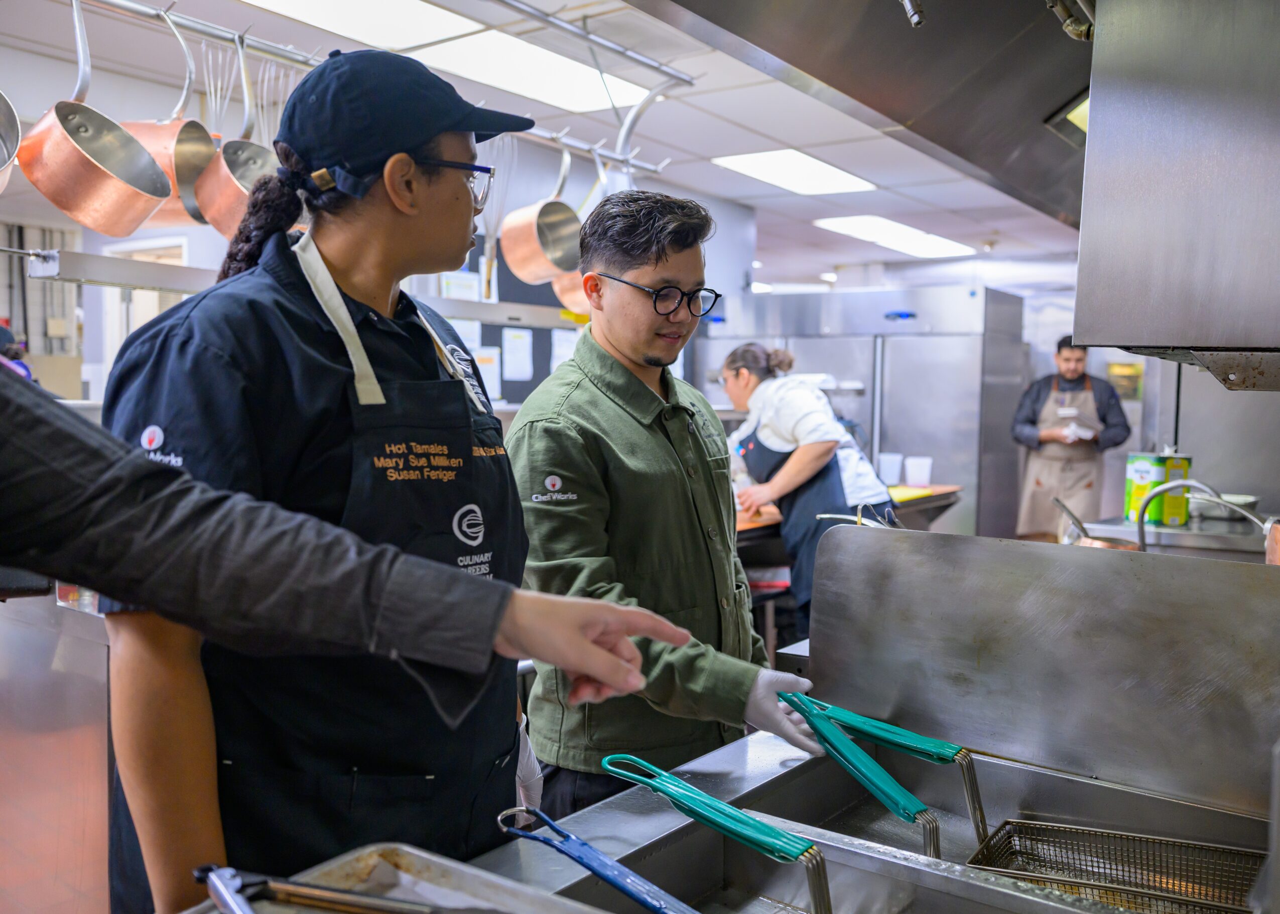 Two people in a kitchen, one wearing a chef's uniform and the other in casual clothing, cooking at a stove.