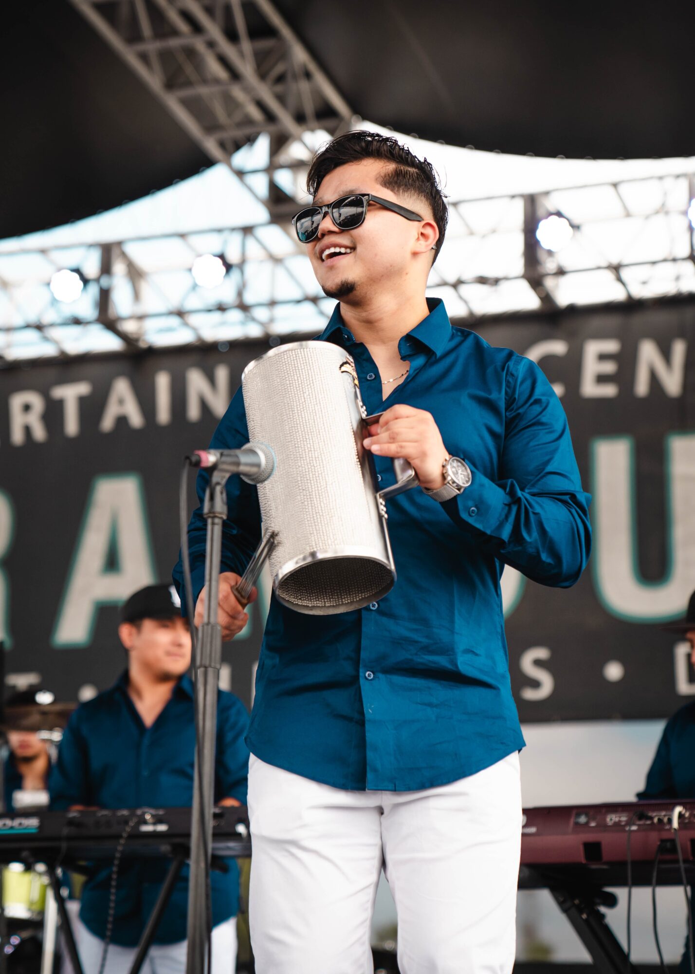 Young man in blue shirt and white pants holding a microphone on stage, smiling, with a band in the background.